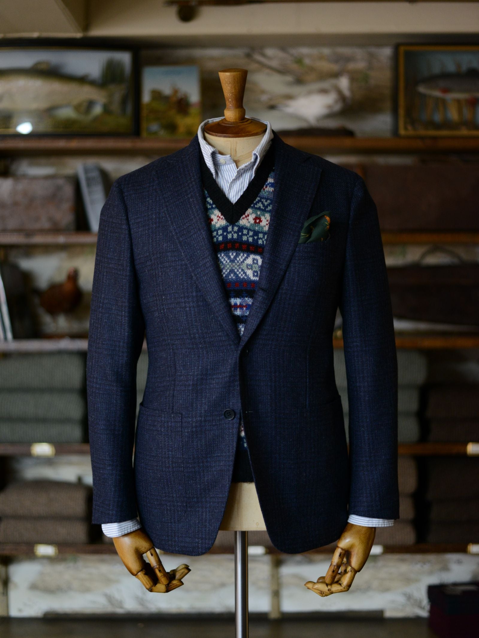 A mannequin dressed in a Campbell's of Beauly Auld Stock Glen Check Jacket, sweater vest, collared shirt, and green pocket square stands in a warmly lit rustic room with shelves and framed pictures in the background.