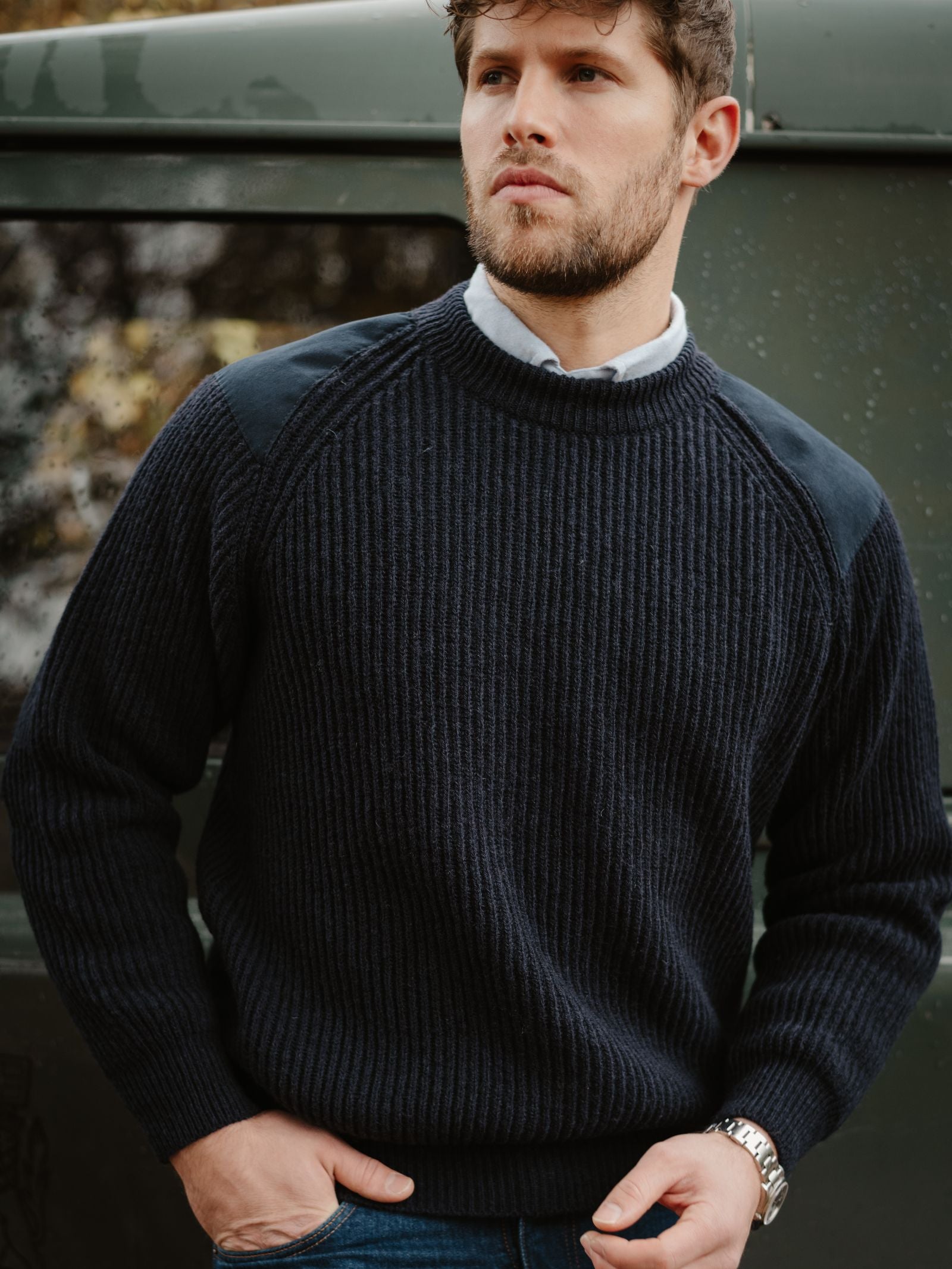 A man with short curly hair and a beard stands by a green vehicle, wearing the Campbell's of Beauly Scotia Suede Jumper over a collared shirt, paired with blue jeans and a wristwatch, looking thoughtfully to the side.