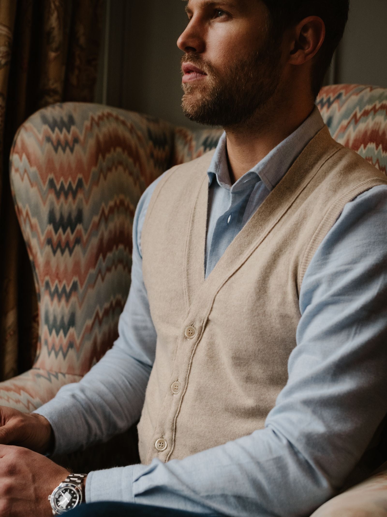 A man wearing a Campbell's of Beauly Geelong Lambswool Button Waistcoat over a blue dress shirt sits in a patterned armchair, looking to the side. He has a short beard, wears a watch on his left wrist, with patterned upholstery and curtains in the background.