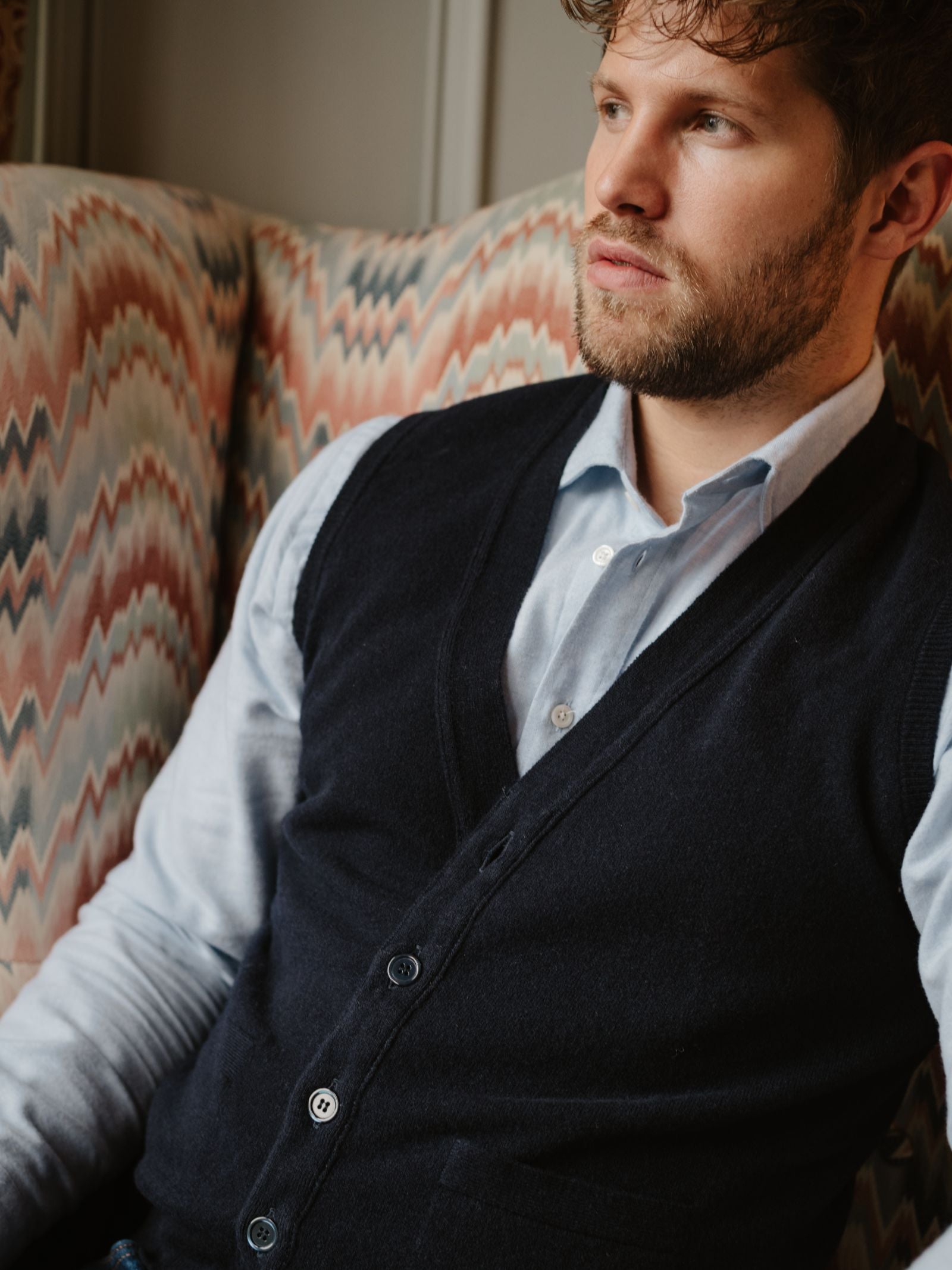 A bearded man in a light blue shirt and a Campbell's of Beauly Geelong Lambswool Button Waistcoat sits on a patterned armchair, gazing thoughtfully to the side.