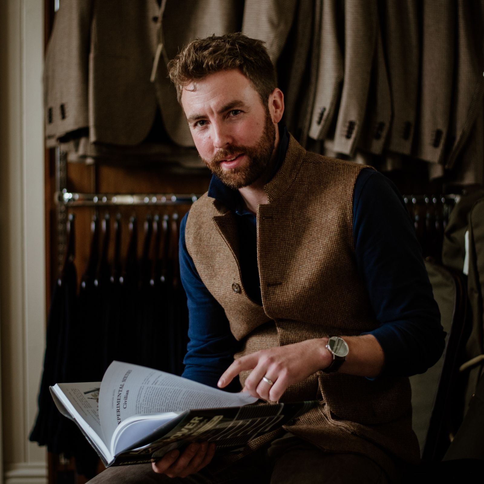 A bearded man in a brown vest sits indoors, holding open a book and smiling slightly while looking at the camera. Behind him are hanging suits and jackets on a rack.