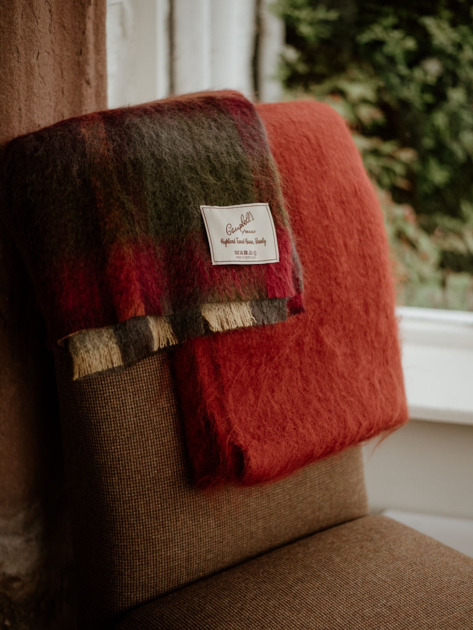 A brown upholstered chair with two folded wool blankets over the back: a red Mohair Throw by Campbells of Beauly, adding a luxurious touch, and a red and green plaid with a Quality Supreme Highland Tweeds label from Scotland.