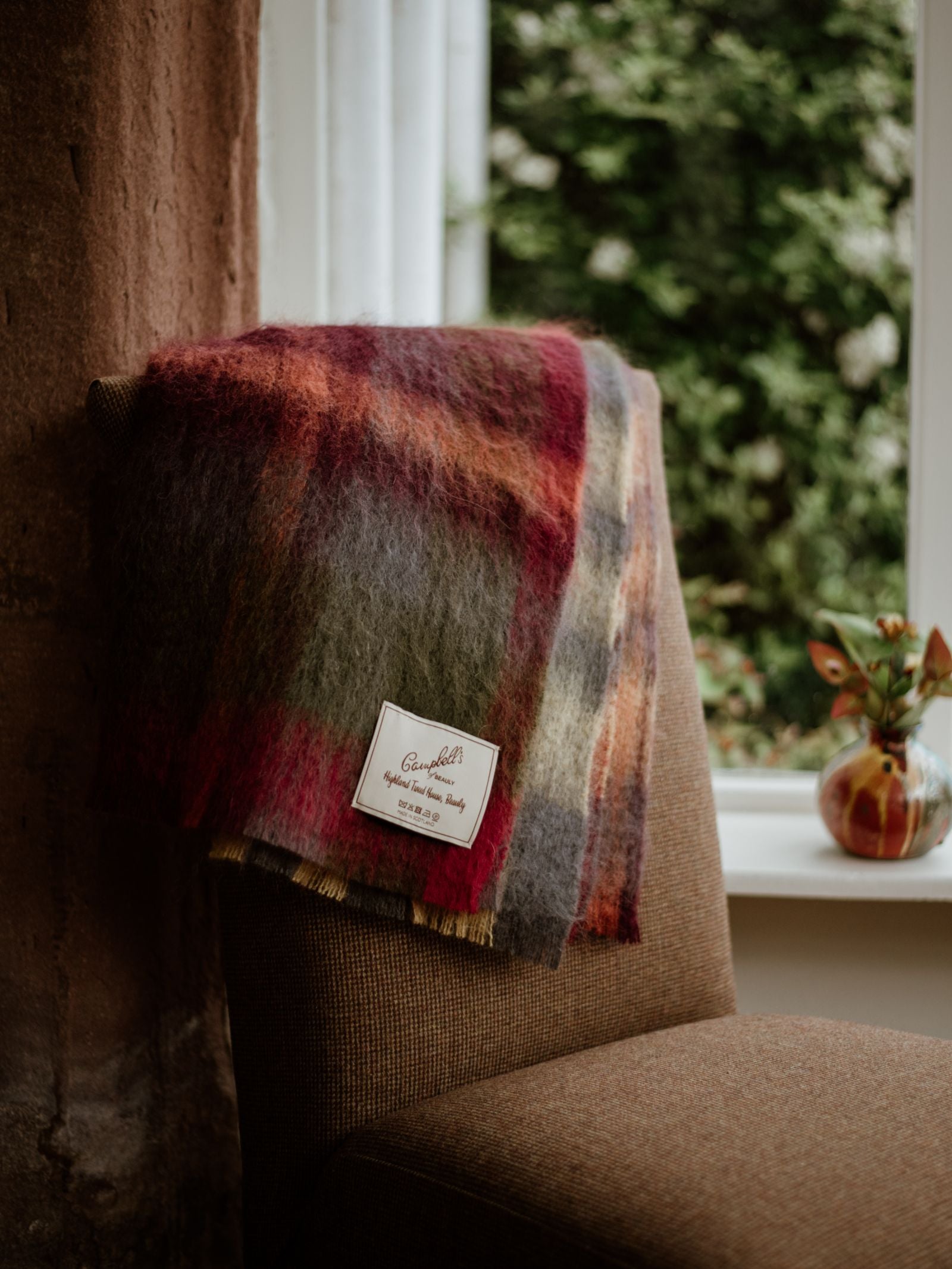 The Campbells of Beauly Painterly Mohair Throw is draped over a brown upholstered chair by a sunlit window, with a small vase of flowers perched on the windowsill beside it.