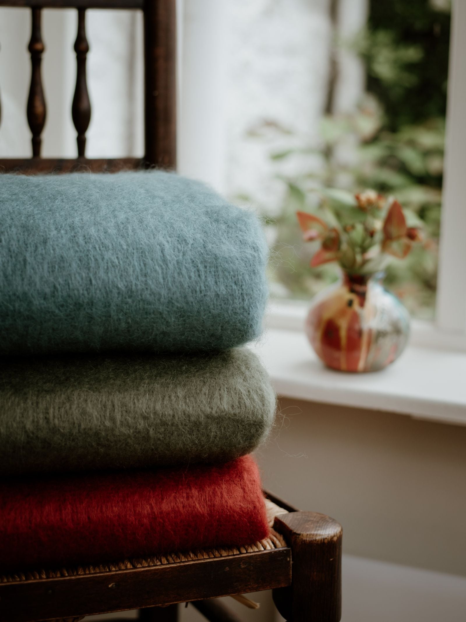 A stack of three Campbells of Beauly Mohair Throws in blue, green, and red rests on a wooden chair by the window. On the sill, a small vase with leaves and flowers is framed by soft Yorkshire light.