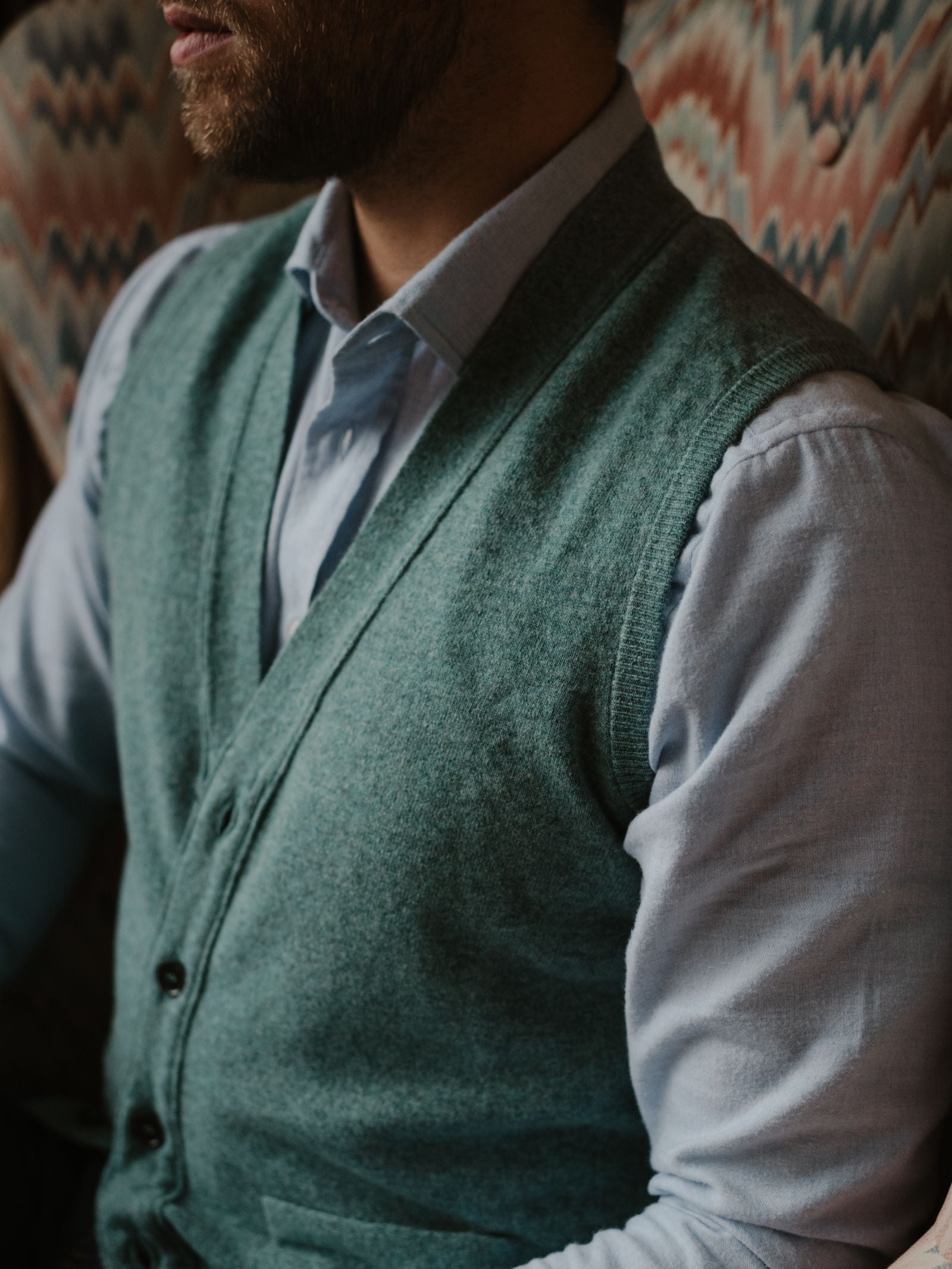 A man in a light blue dress shirt and a green Campbell's of Beauly Geelong Lambswool Button Waistcoat sits on a patterned chair, his classic country wardrobe the focal point.