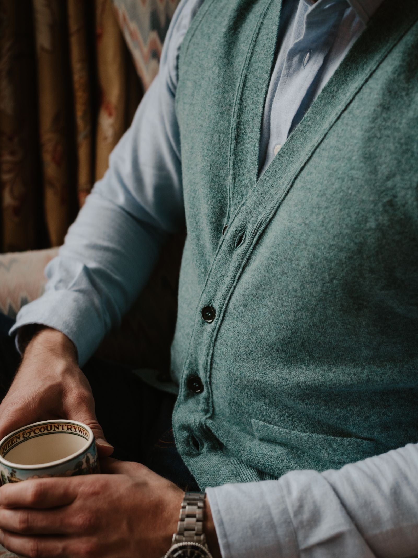A person in a light blue shirt and a green Campbell's of Beauly Geelong Lambswool Button Waistcoat holds a patterned teacup. Only their torso and hands are shown, with a watch on the left wrist and country wardrobe upholstery in the background.