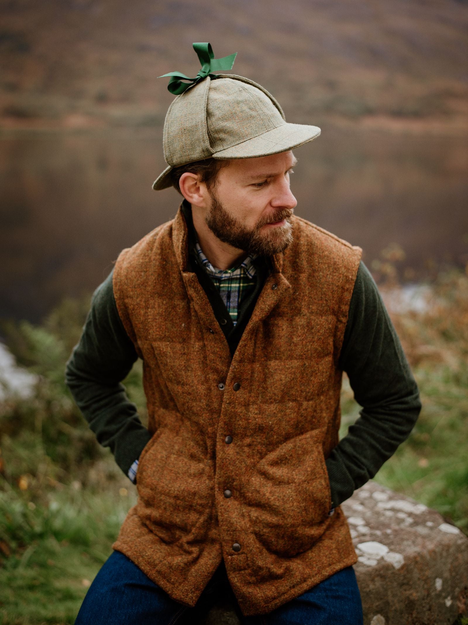 A bearded man in a Campbell's of Beauly Harris Tweed Quilted Gilet, green plaid shirt, and green jacket sits outdoors on a rock, wearing a deerstalker hat with a green ribbon, gazing at a lake and hills in the background.