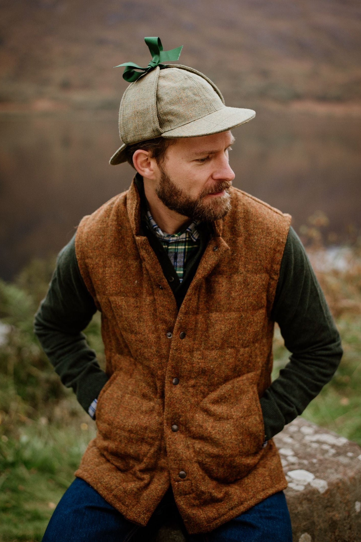 A bearded man in a deerstalker hat, plaid shirt, and Campbell's of Beauly Harris Tweed Quilted Gilet sits outdoors with hands in pockets, glancing sideways against a blurred natural background.