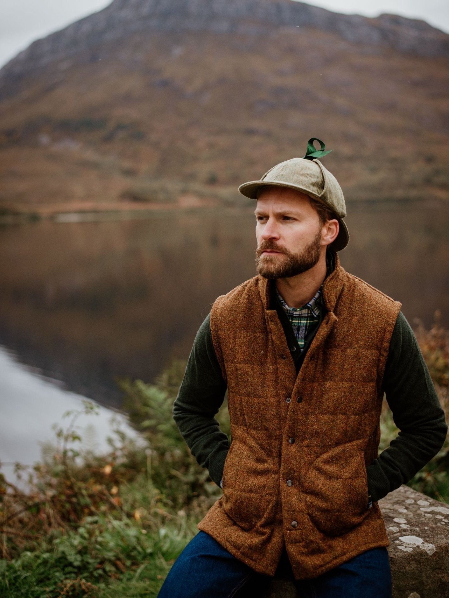 A bearded man in a Campbell's of Beauly Harris Tweed Quilted Gilet, plaid shirt, and cap sits by a calm lake with autumn trees and a mountain in the background.