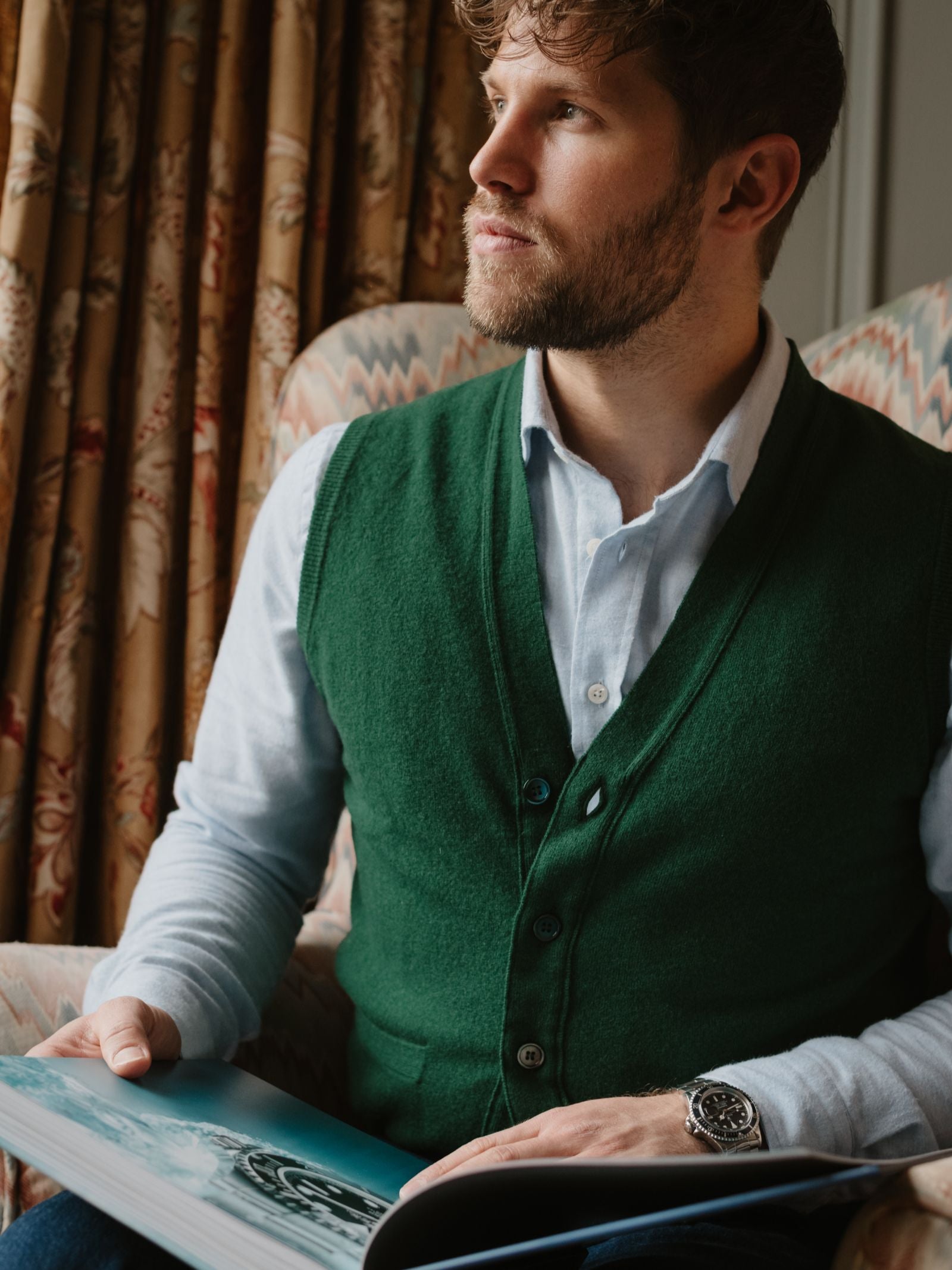 A bearded man in a green Campbell's of Beauly Geelong Lambswool Button Waistcoat and light blue shirt sits in a patterned chair, holding an open book as window light highlights the timeless charm of classic country style.
