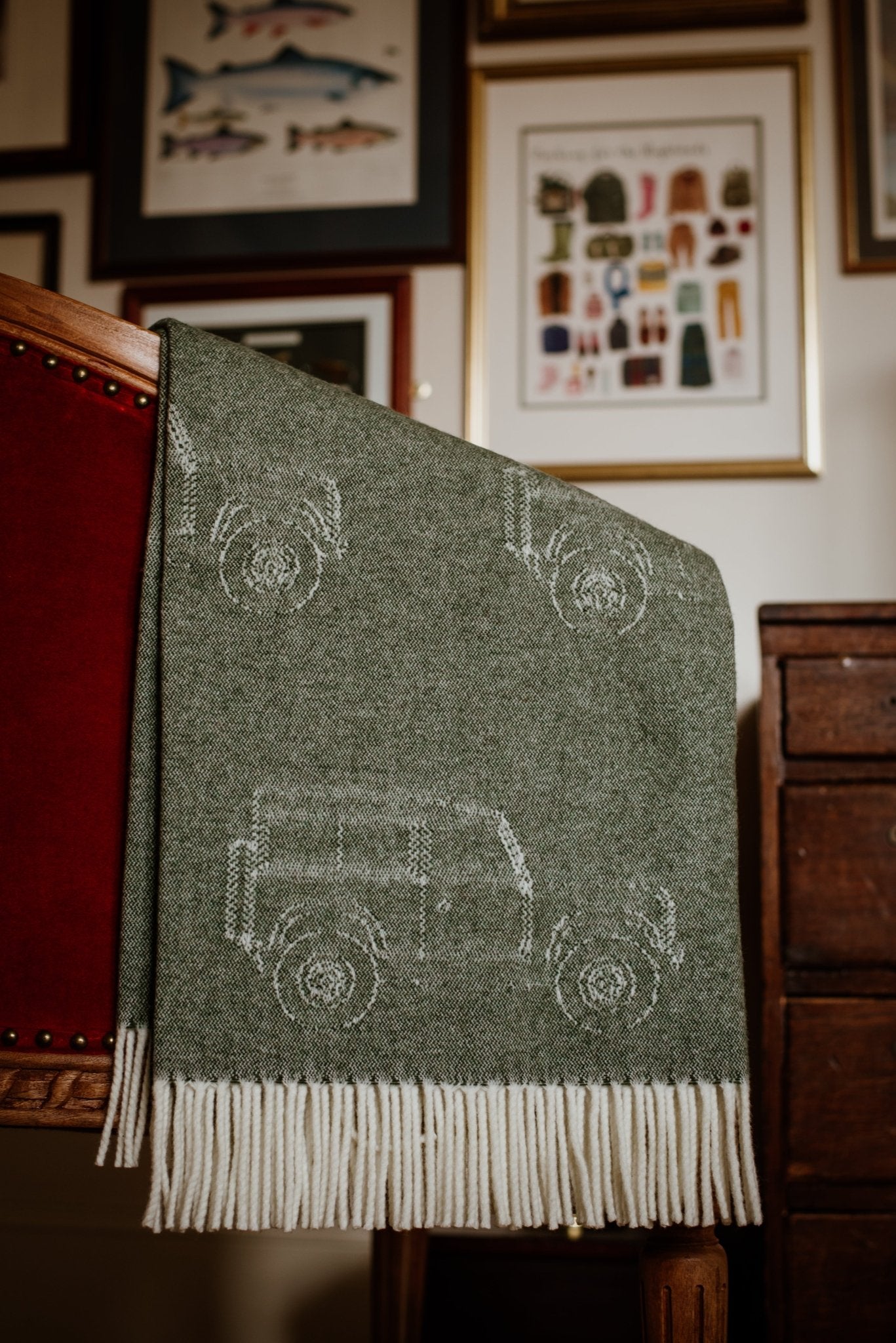 The Land Rover Defender Jacquard Throw by Campbells of Beauly, a green wool blanket with white fringe and off-road vehicle motifs, is draped over a red upholstered wooden chair. Framed artwork and drawings appear on the wall behind it.