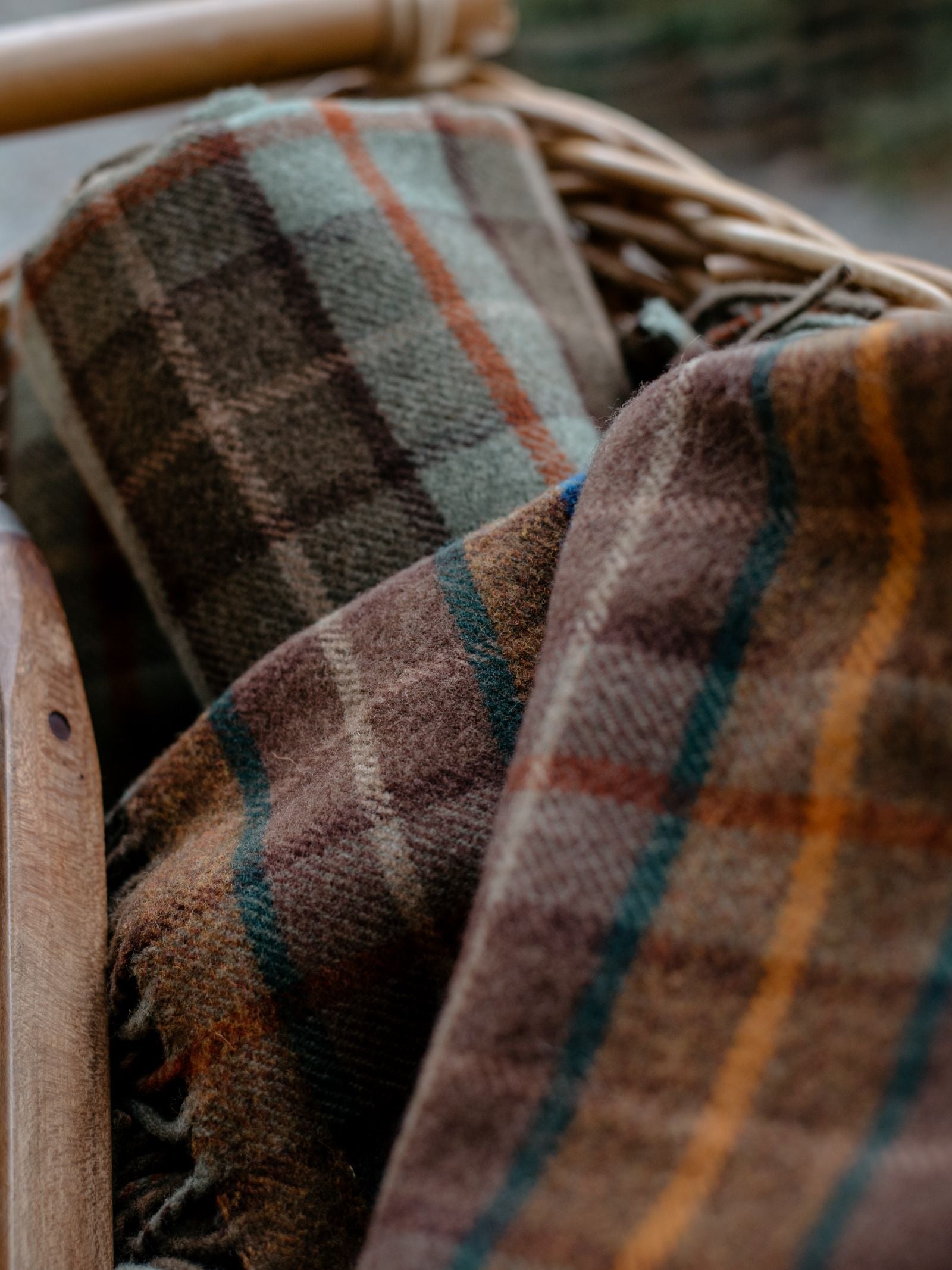 Close-up of two Campbells of Beauly Lavender Throws with plaid patterns in earthy colors, draped over a wicker basket—crafted with care in the Scottish Borders.
