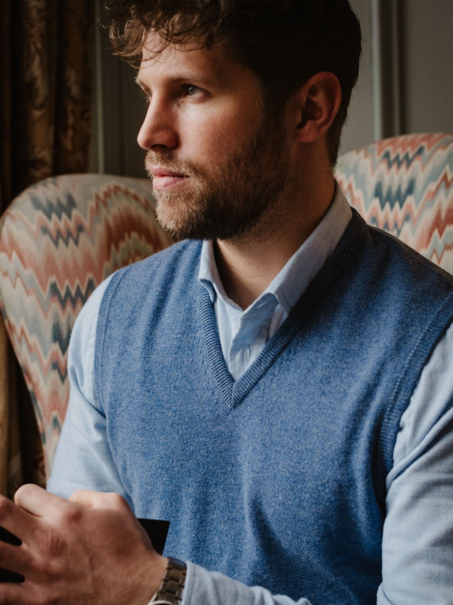A man with a beard and wavy hair sits indoors, thoughtfully looking to the side while wearing the Campbell's of Beauly Cashmere Slipover in light blue. He holds a dark object, with patterned chairs visible in the background.