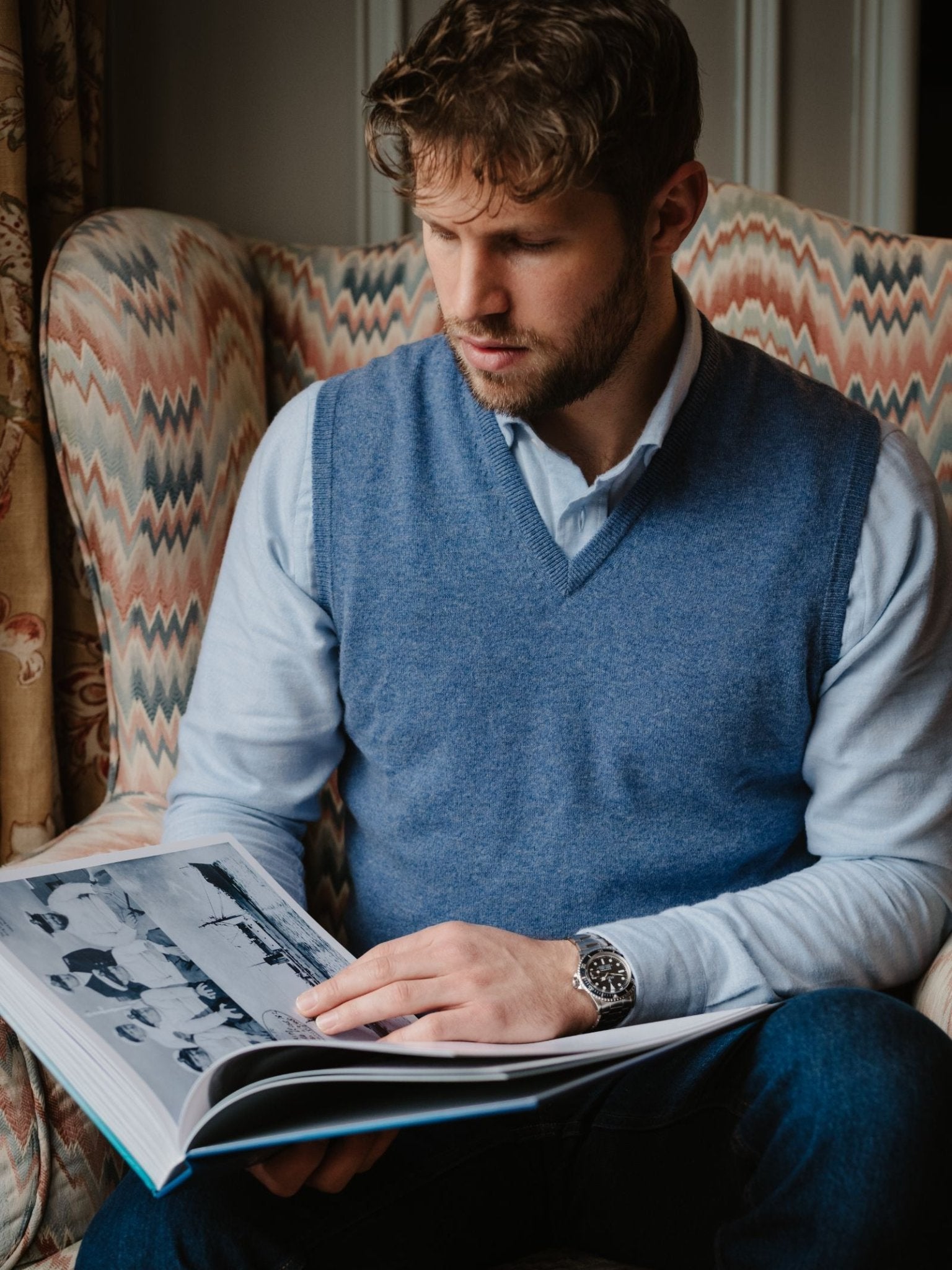 A man with wavy brown hair and a trimmed beard sits in an armchair, wearing the Campbell's of Beauly Cashmere Slipover over a light shirt, looking at a large open photo book with black-and-white images.