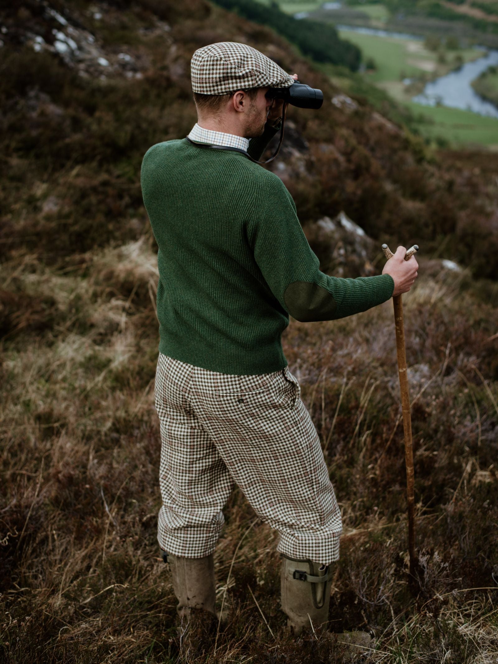 Wearing a Campbells of Beauly Fishermans Rib Crew Jumper, a man with suede elbow patches, plaid cap, and checked trousers stands on a hillside with a walking stick, scanning the landscape below through binoculars.
