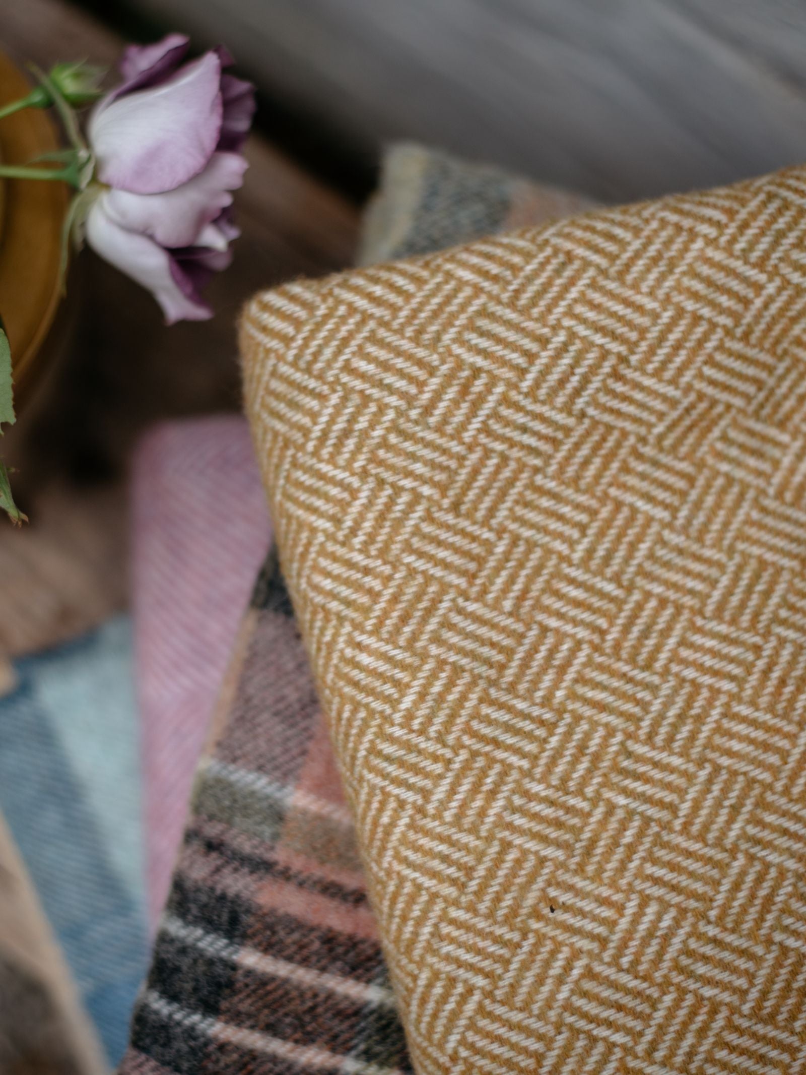 A close-up of Campbells of Beaulys Parquet Lambswool Blanket showcases folded woven fabrics in earth tones with parquet, plaid, and striped patterns. A mauve rose in the top left adds softness to the Yorkshire-inspired details.