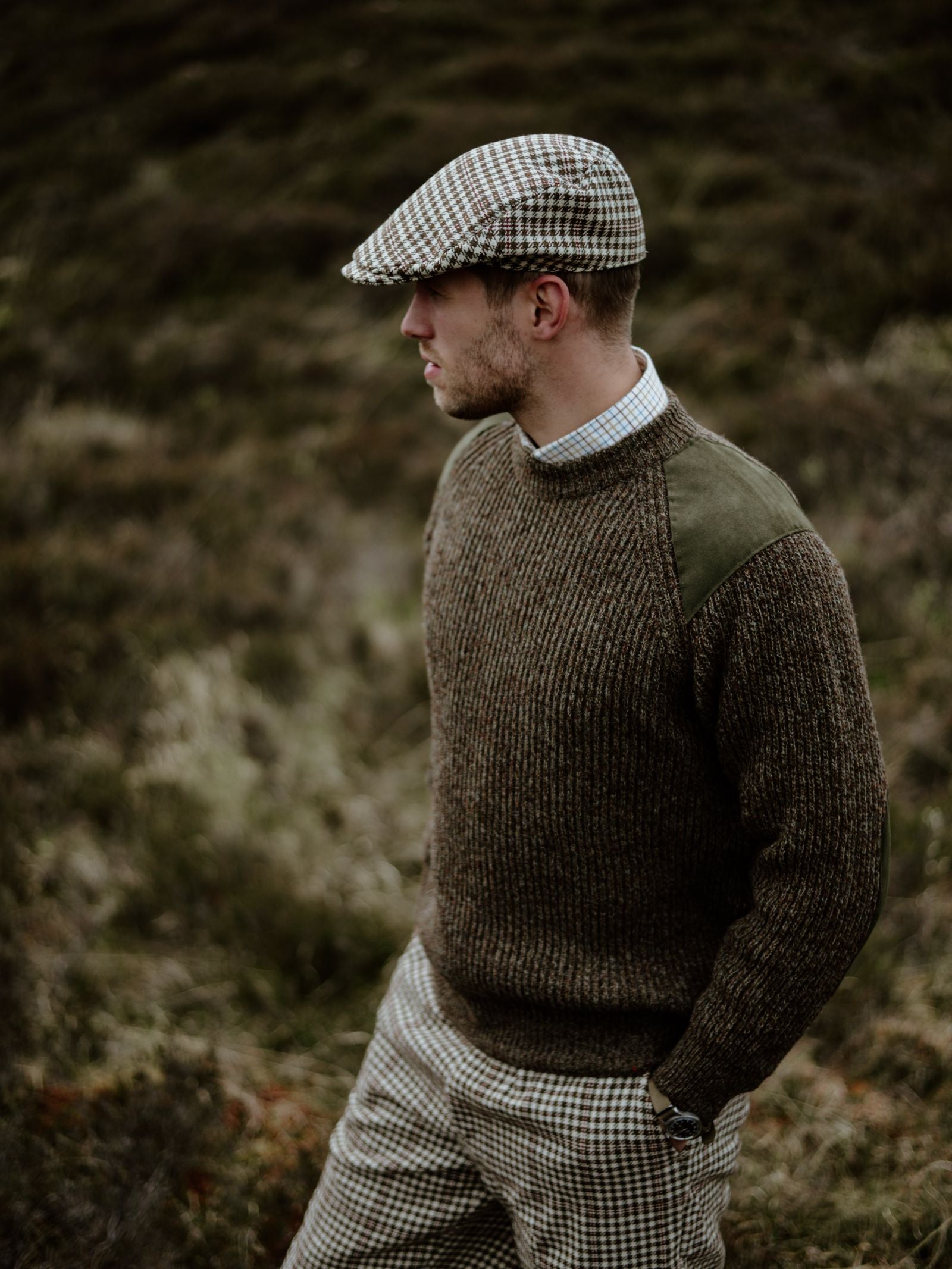 A man in Campbells of Beauly Scotia Suede Jumper with suede shoulder patches, a checked flat cap, and checked trousers stands on grassy terrain, gazing to the side with one hand in his pocket.