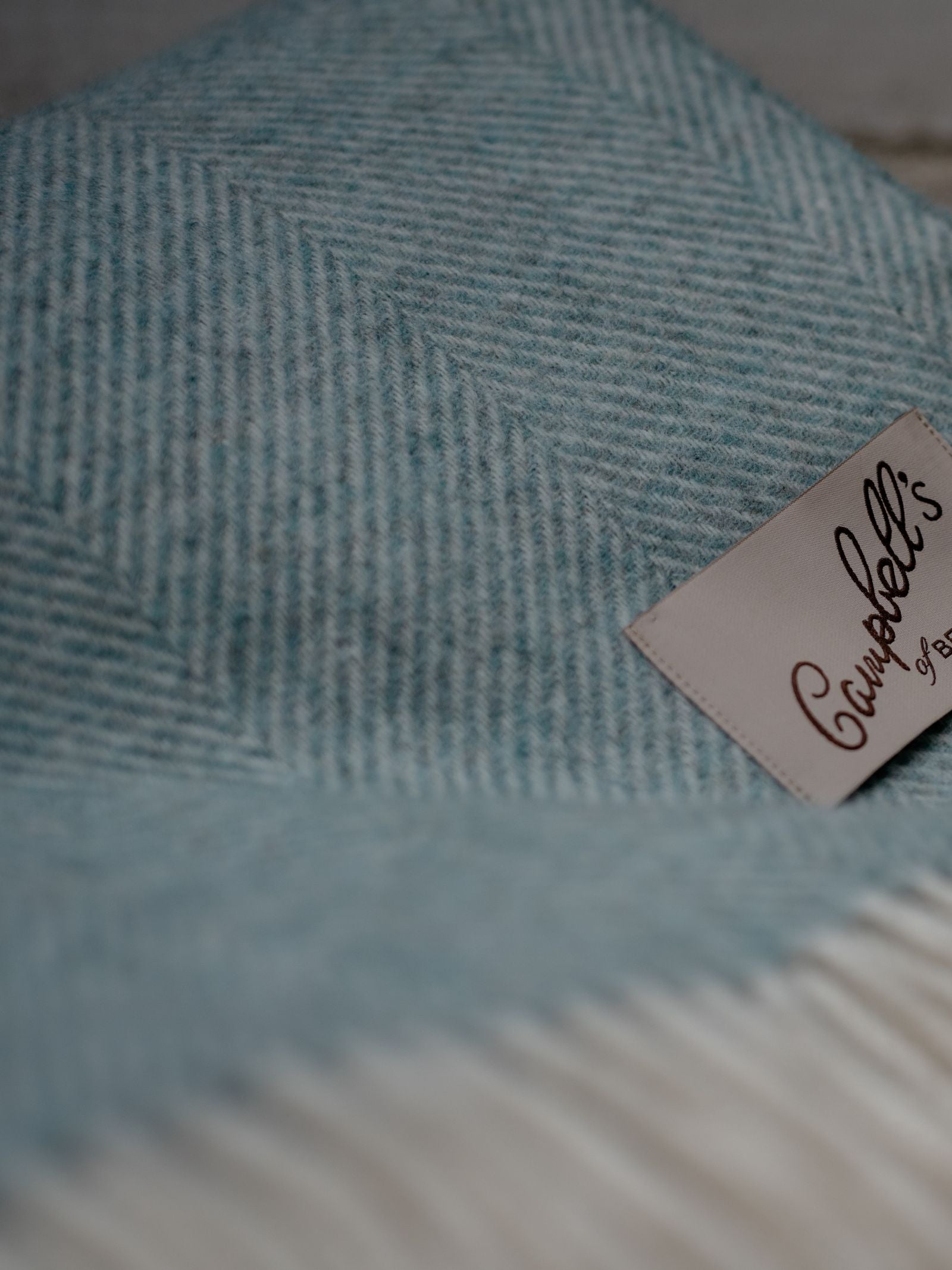 A close-up of the Campbells of Beauly Herringbone Lambswool Blanket in duck egg, showing its soft texture and signature beige tag.