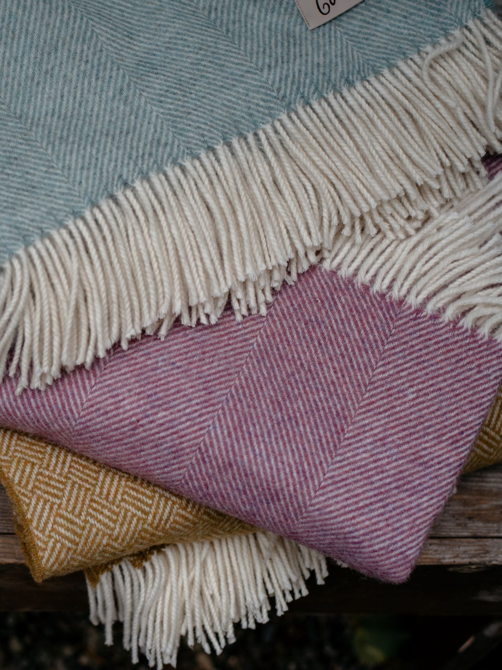 A Campbells of Beauly Herringbone Lambswool Blanket in pink with white fringes is neatly folded and stacked on a wooden surface, showcasing its classic woven pattern and soft texture.