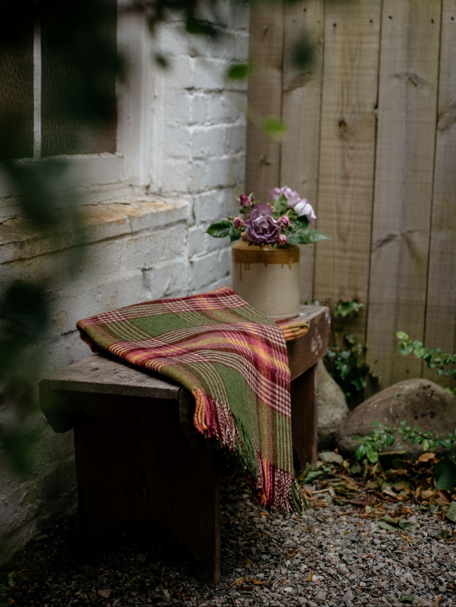 A wooden bench, draped with the Campbells of Beauly Kilmorack Lambswool Throw in a green and red check, sits by a white brick wall. A ceramic pot of purple flowers rests on the bench. Gravel and stones cover the ground near a wooden fence.
