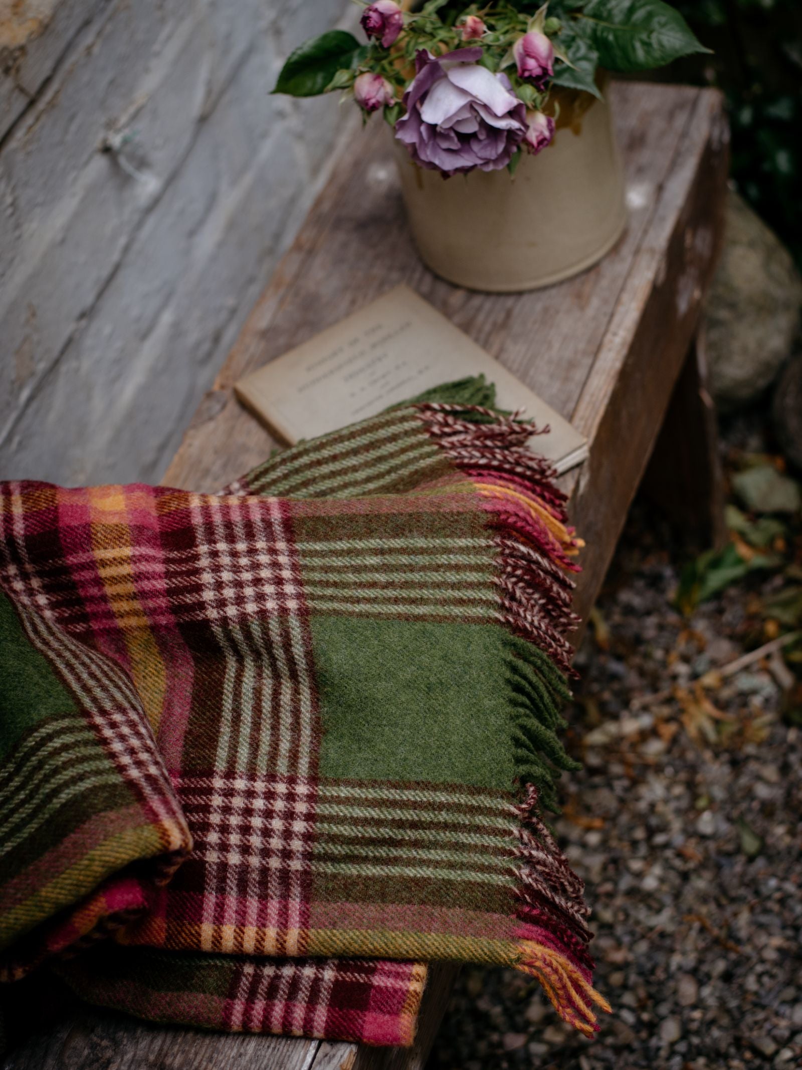 A folded Campbells of Beauly Kilmorack Lambswool Throw in a classic green, red, and brown check rests on a rustic wooden bench beside an old book and purple flowers, with gravel and stones nearby.
