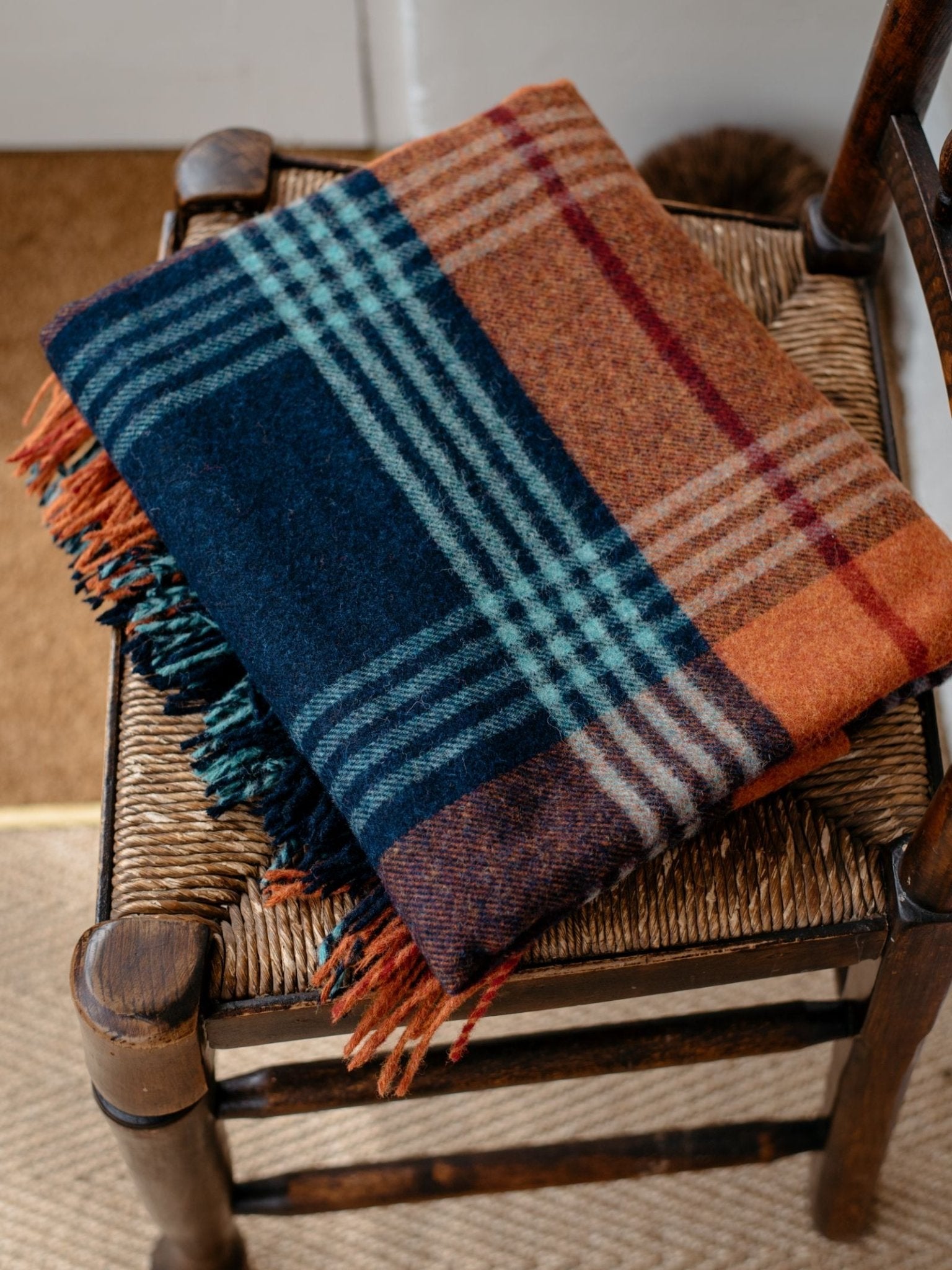 The Campbells of Beauly Torgormack Lambswool Throw, in blue, orange, brown, and cream plaid with fringed edges, sits folded on a rustic woven chair, bringing cosy Yorkshire-inspired style to the space.