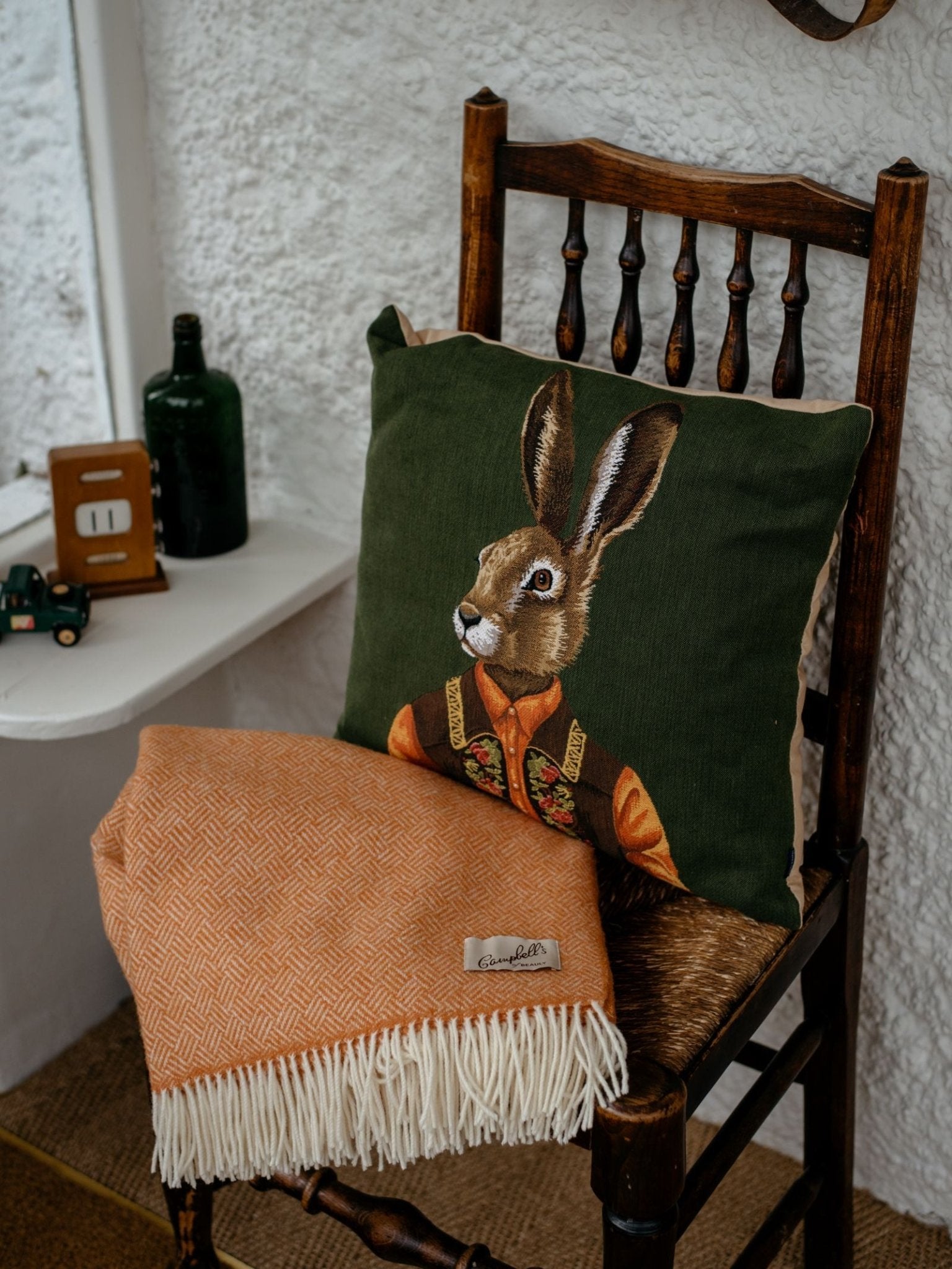 A wooden chair displays the Parquet Lambswool Blanket by Campbells of Beauly in orange with fringe, along with a rabbit-themed cushion; beside it, a small white table holds a green bottle and toy tractor against a textured white wall.