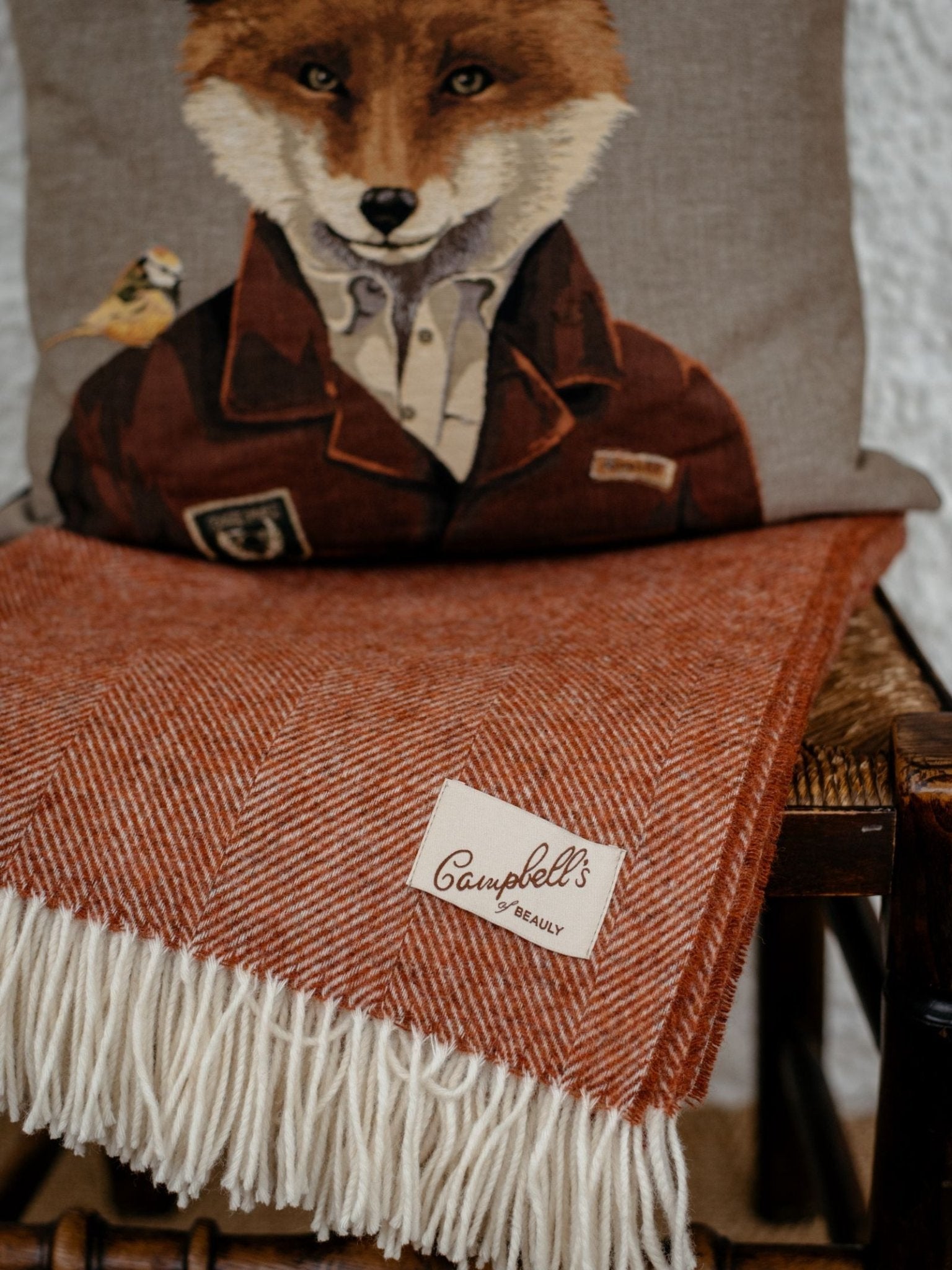 A reddish-brown Campbells of Beauly Herringbone Lambswool Blanket with white fringe is draped over a chair. Behind it sits a cushion featuring an illustrated fox in a suit jacket and shirt.