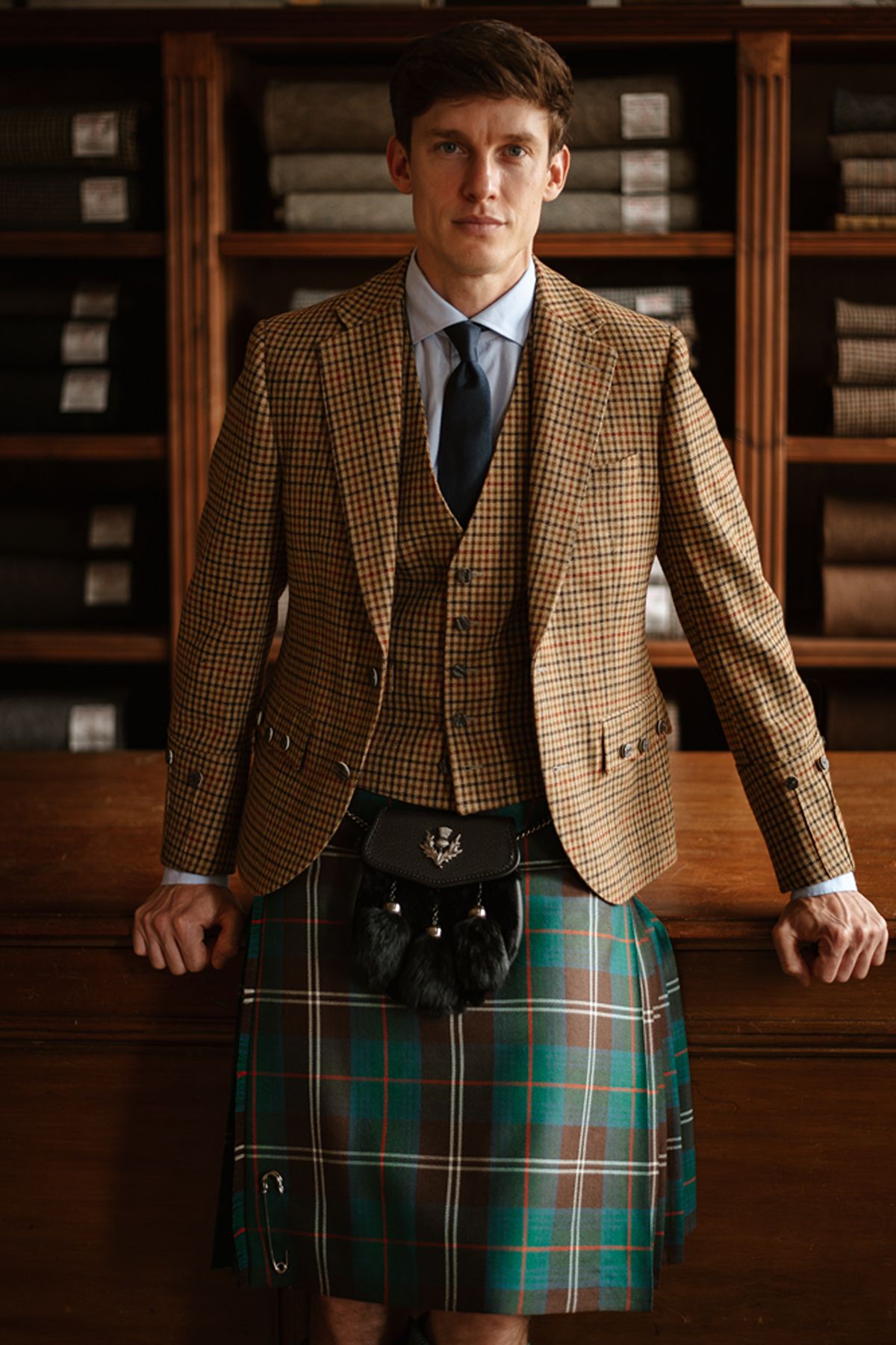 A man wearing a tweed jacket, waistcoat, and tartan kilt with a leather sporran stands before shelves of fabric bolts.