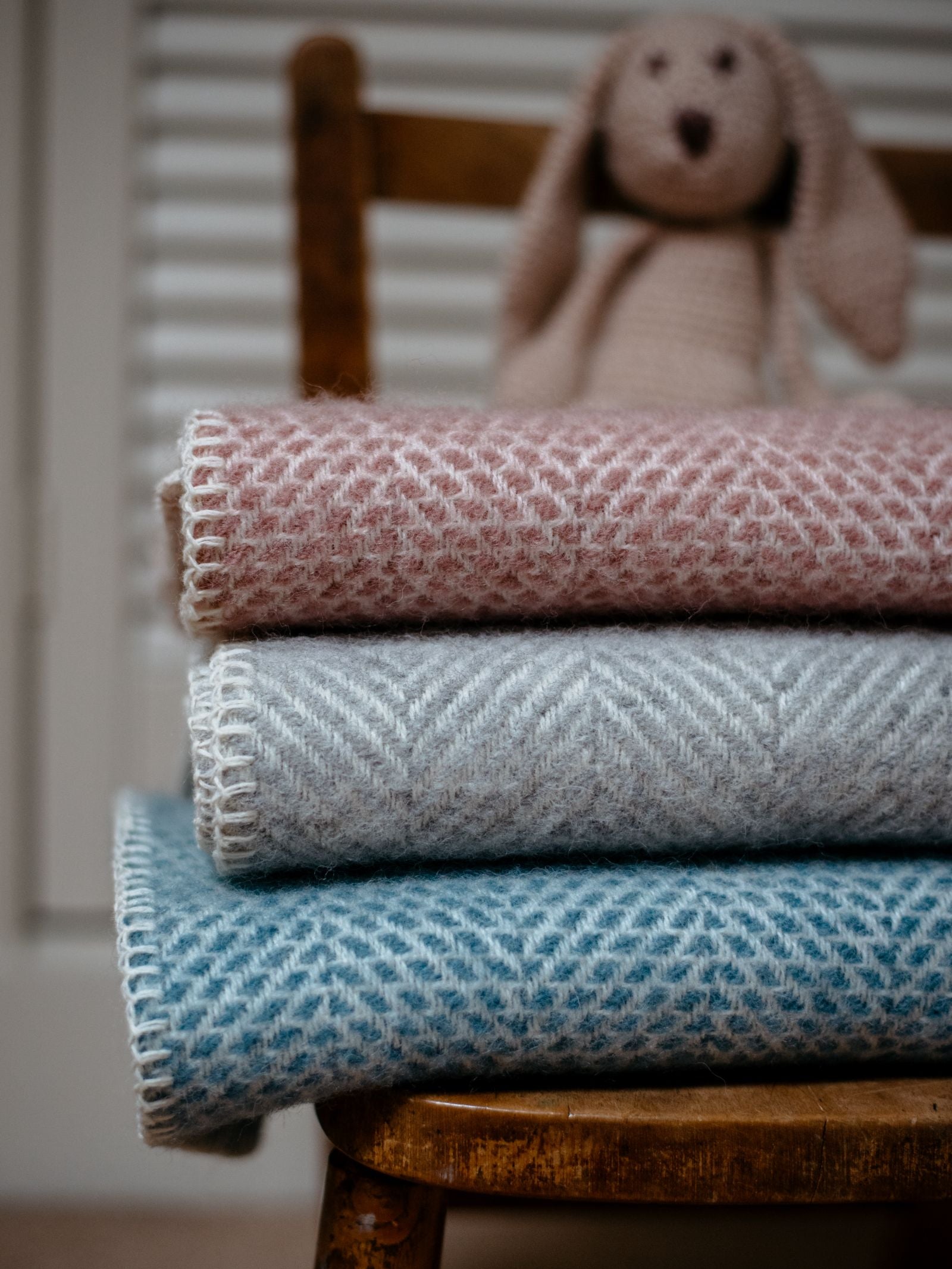 Three Campbells of Beauly Baby Blankets in pink, gray, and blue are neatly folded on a wooden chair, providing year-round comfort. A knitted bunny toy sits in the background.