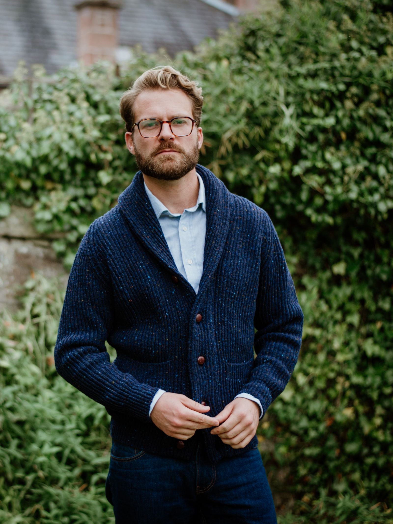 A man with light brown hair, beard, and glasses stands outdoors amid greenery, wearing Campbell's of Beauly Donegal Lambswool Shawl Collar Cardigan in blue over a light blue shirt and dark jeans, looking seriously at the camera.