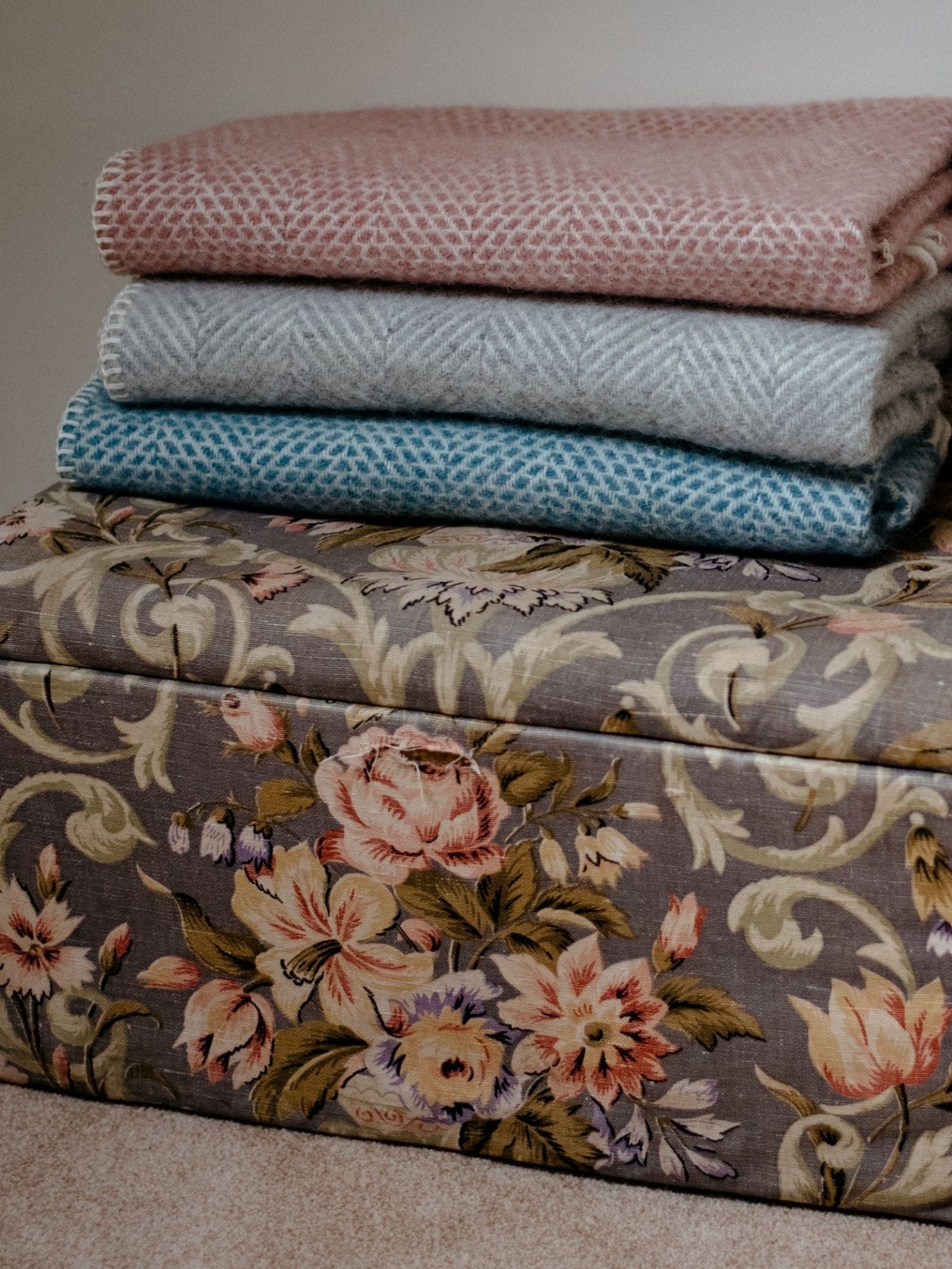 Three Campbells of Beauly Baby Blankets in pink, gray, and blue are folded atop a floral storage chest, showcasing their soft weave and muted tones—ideal for cozy comfort in any season.