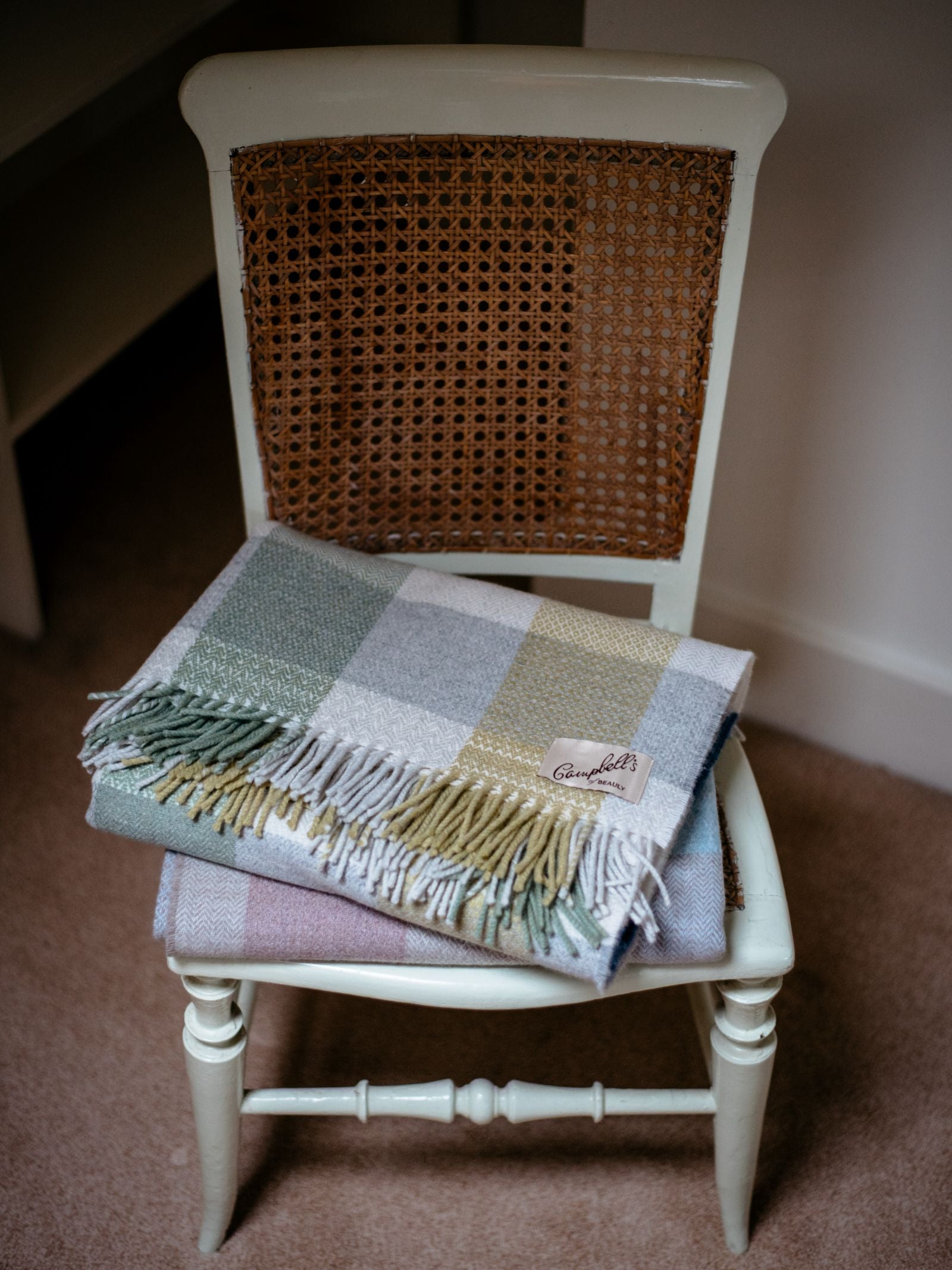 A Campbells of Beauly Patchwork Throw is neatly folded on a vintage wooden chair with a woven cane backrest, which stands on a beige carpet in a gently lit room.
