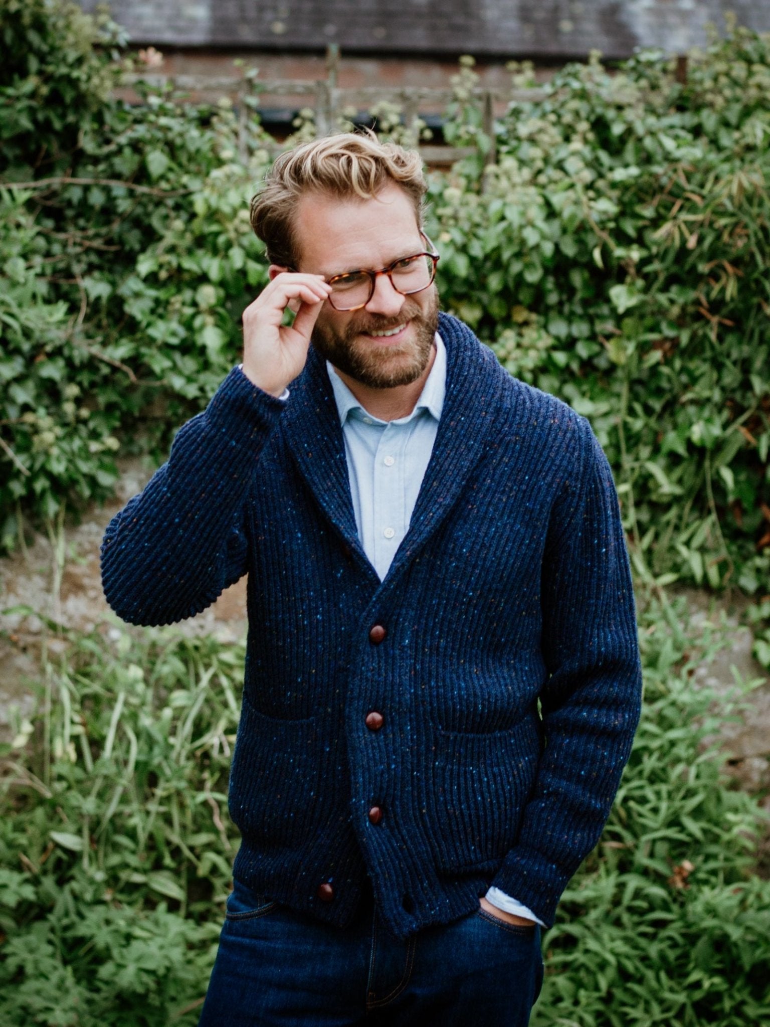 A bearded man with glasses stands outdoors in front of green foliage, smiling as he touches his glasses. He wears a Donegal Lambswool Shawl Collar Cardigan by Campbell's of Beauly, a light blue shirt, and jeans.
