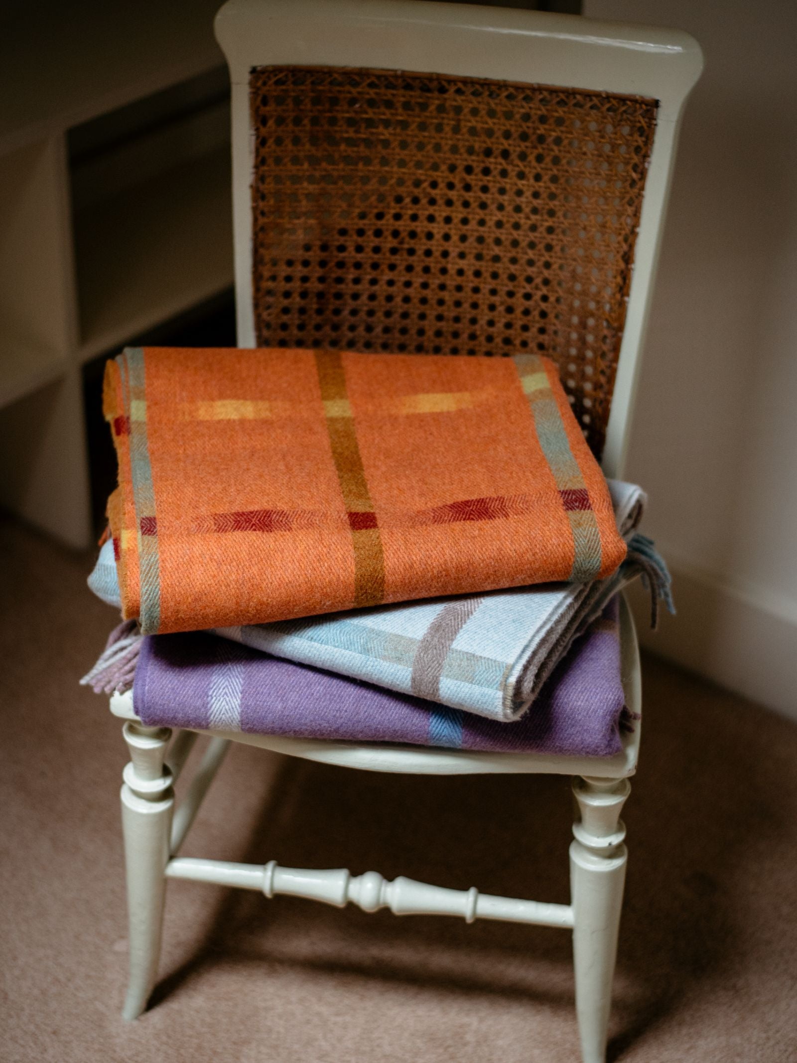 A white wooden chair with a woven cane backrest holds two blankets: an orange windowpane one on top and the Maree Lambswool Blanket by Campbells of Beauly, featuring blue and purple stripes, underneath, set on a carpeted indoor floor.
