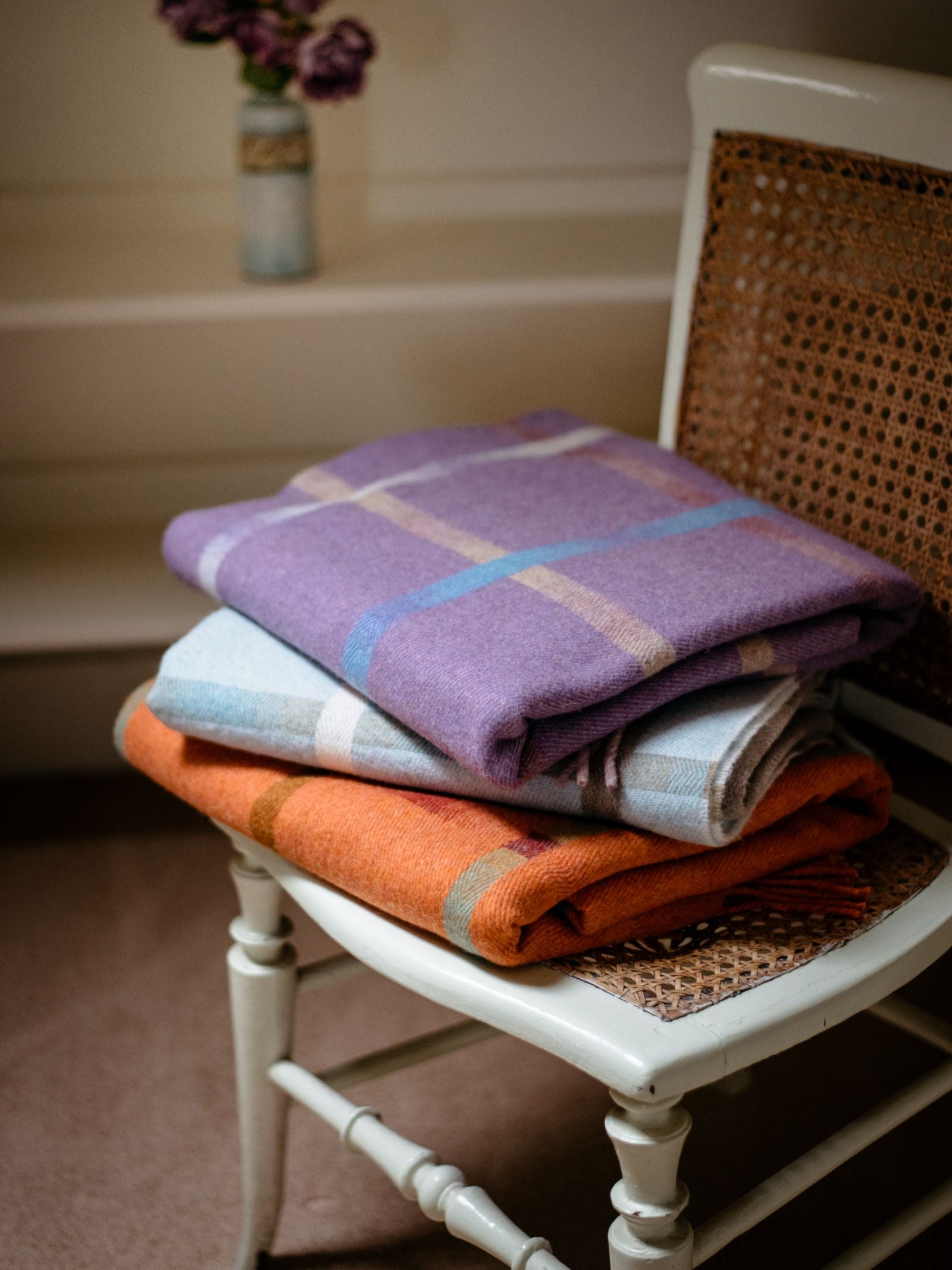 Two folded Maree Lambswool Blankets from Campbells of Beauly, in purple and orange windowpane designs, are stacked on a white vintage cane-back chair. In the background, a blurred vase with purple flowers is visible.