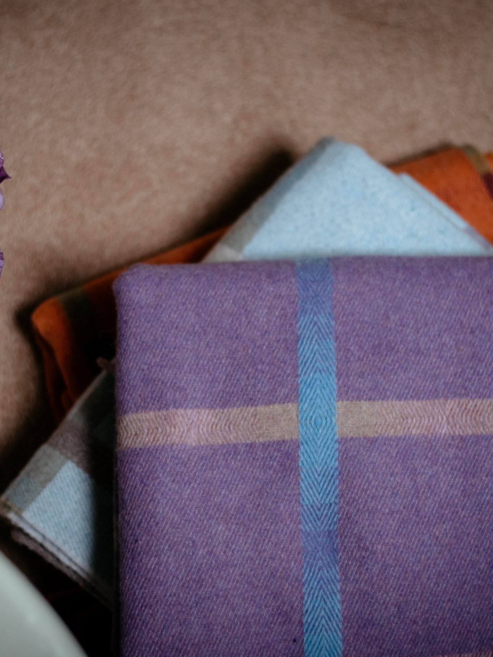 A close-up of folded Maree Lambswool Blankets by Campbells of Beauly, with the top Scottish-made piece in purple with blue windowpane stripes, layered over light blue and orange-toned blankets.