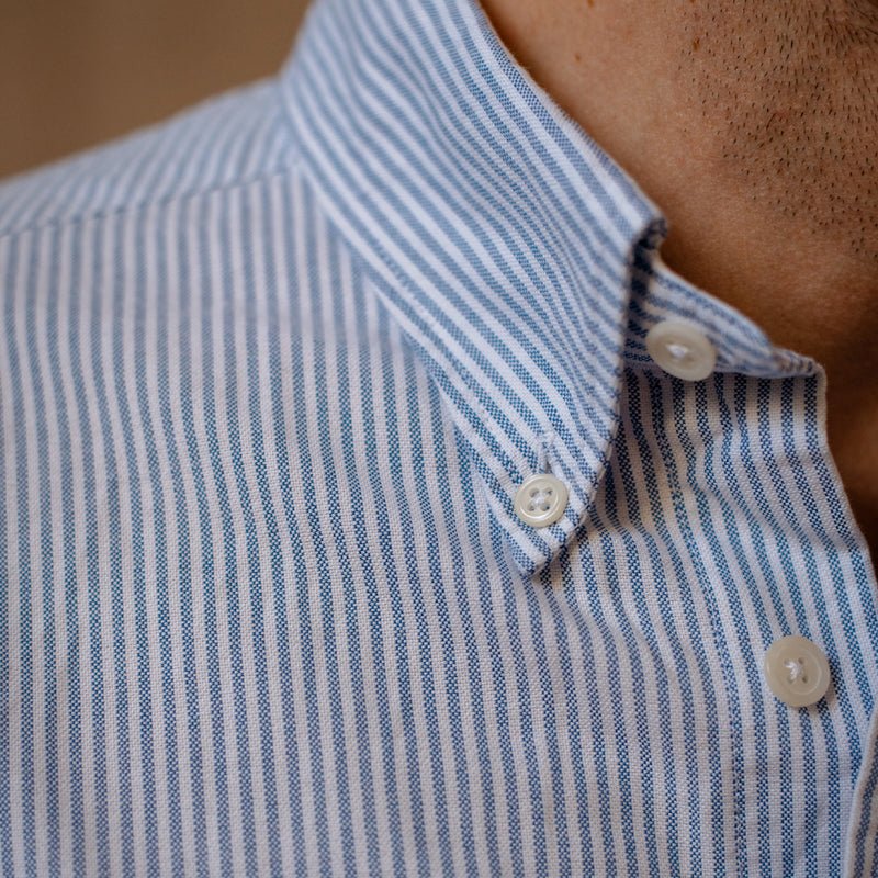Close-up of a persons neck and shoulder wearing the Campbells of Beauly Oxford Shirt in light blue and white stripes, featuring white buttons and a crisp collar. Stubble is visible above the collar.