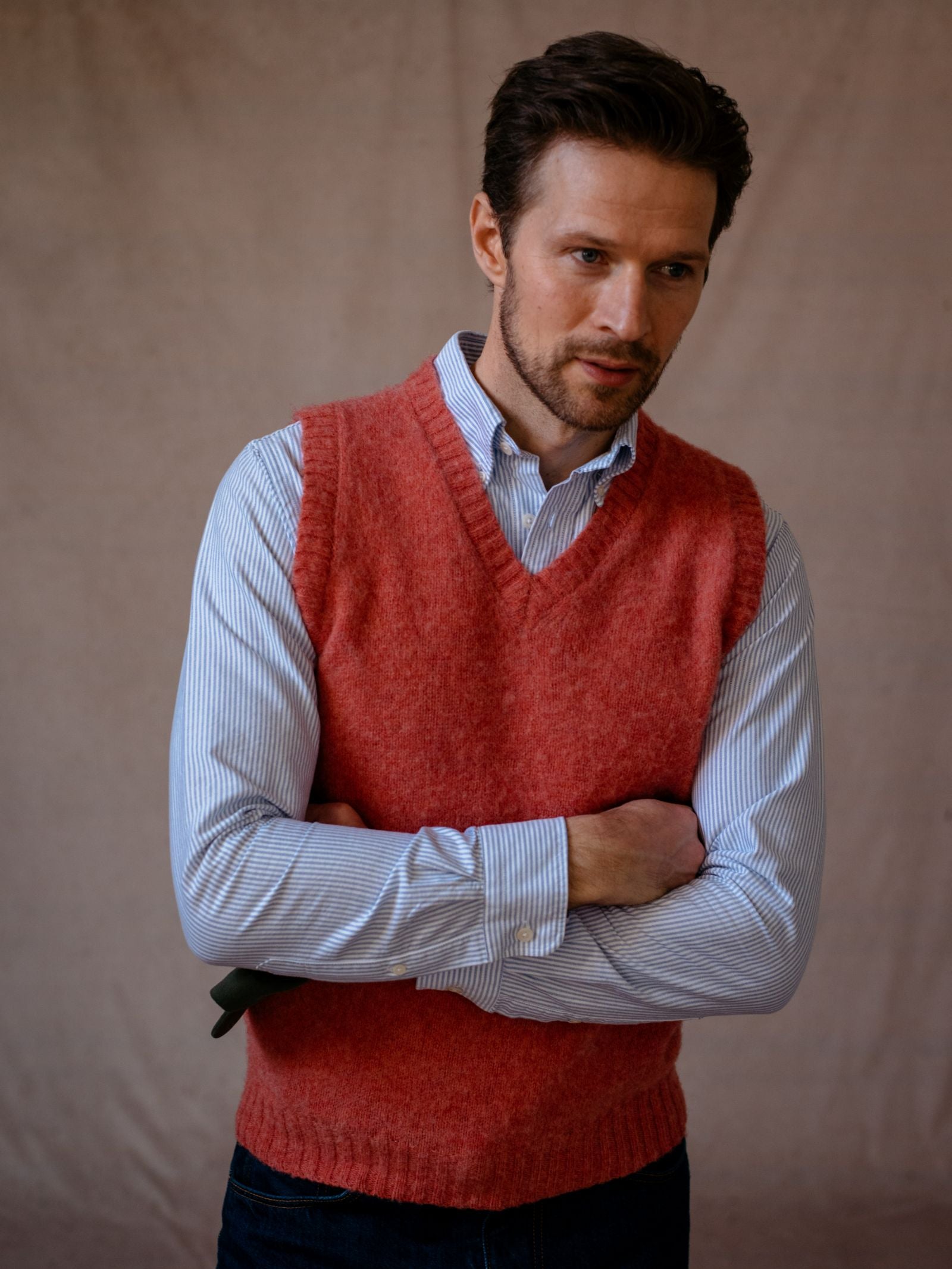 A man with short brown hair and a trimmed beard wears the Campbells of Beauly Shetland Slipover—a red V-neck knit—over a light blue striped dress shirt, standing with arms crossed against a plain beige background.