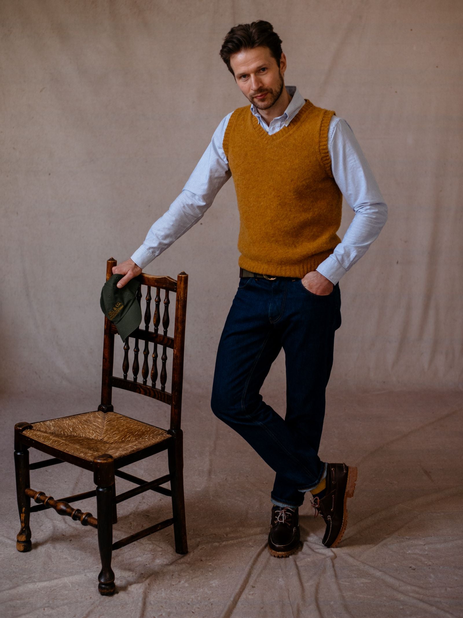 A man wears a Campbells of Beauly Shetland Slipover in mustard yellow over a light blue shirt and dark jeans, standing with one hand in his pocket and leaning on a wooden chair on a light-colored floor.