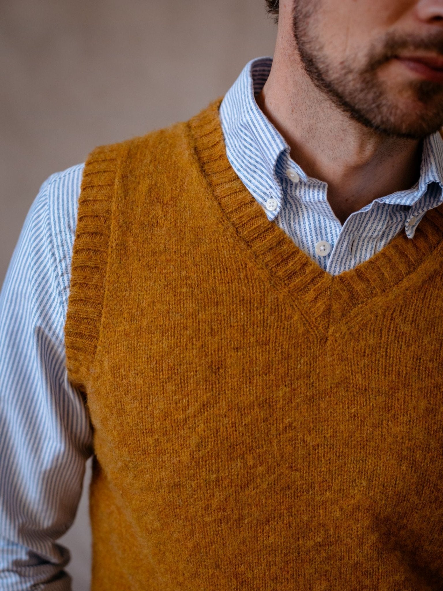 A man wears the Campbells of Beauly Shetland Slipover, a mustard yellow V-neck knit vest, over a blue and white striped shirt. The image highlights this classic piece by showing only the lower face and upper torso.