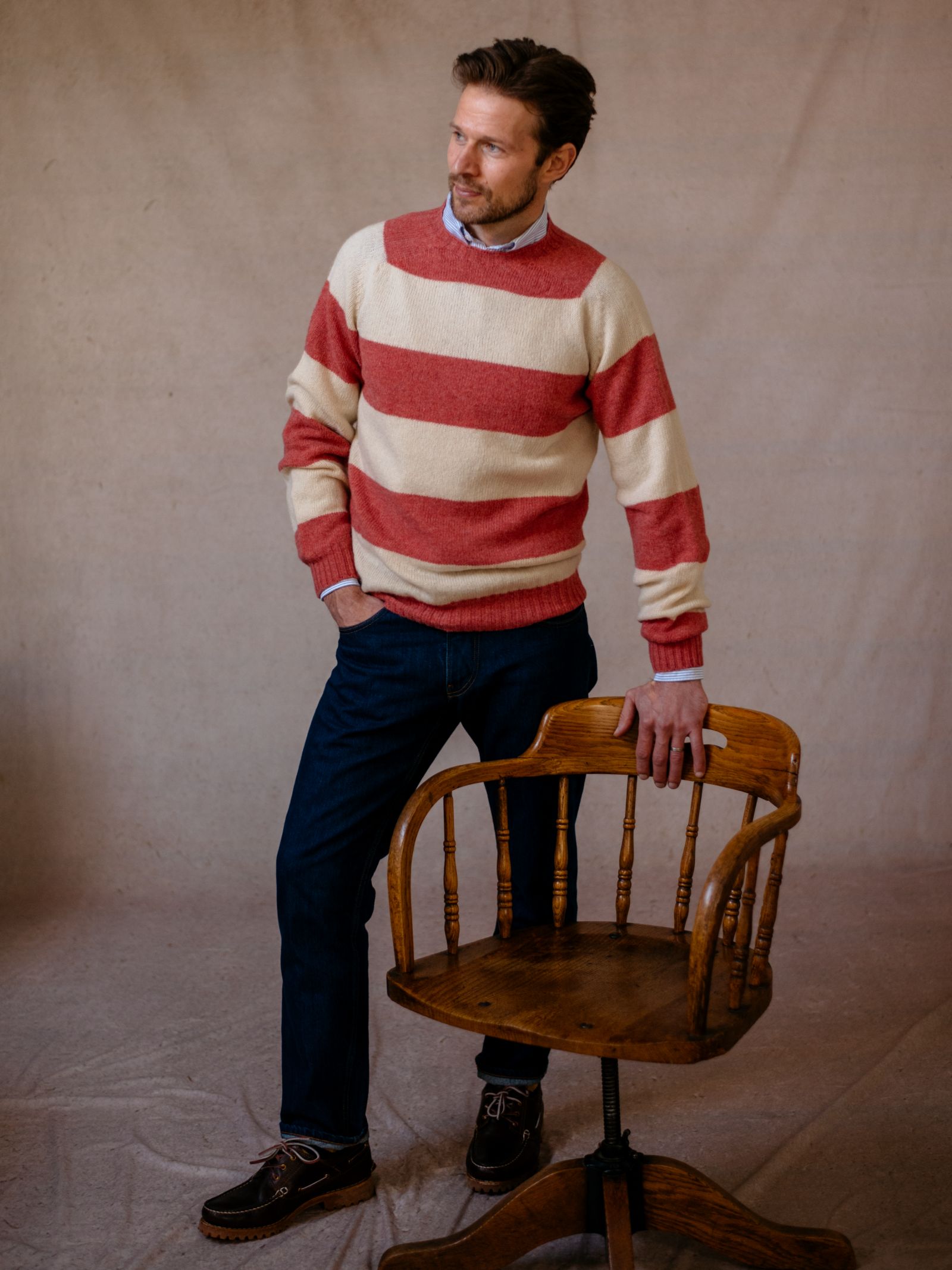 A man stands next to a wooden chair against a beige background, wearing Campbells of Beaulys Shetland Stripe Crew Jumper in red and cream, blue jeans, and brown shoes, with one hand in his pocket and looking to the side.