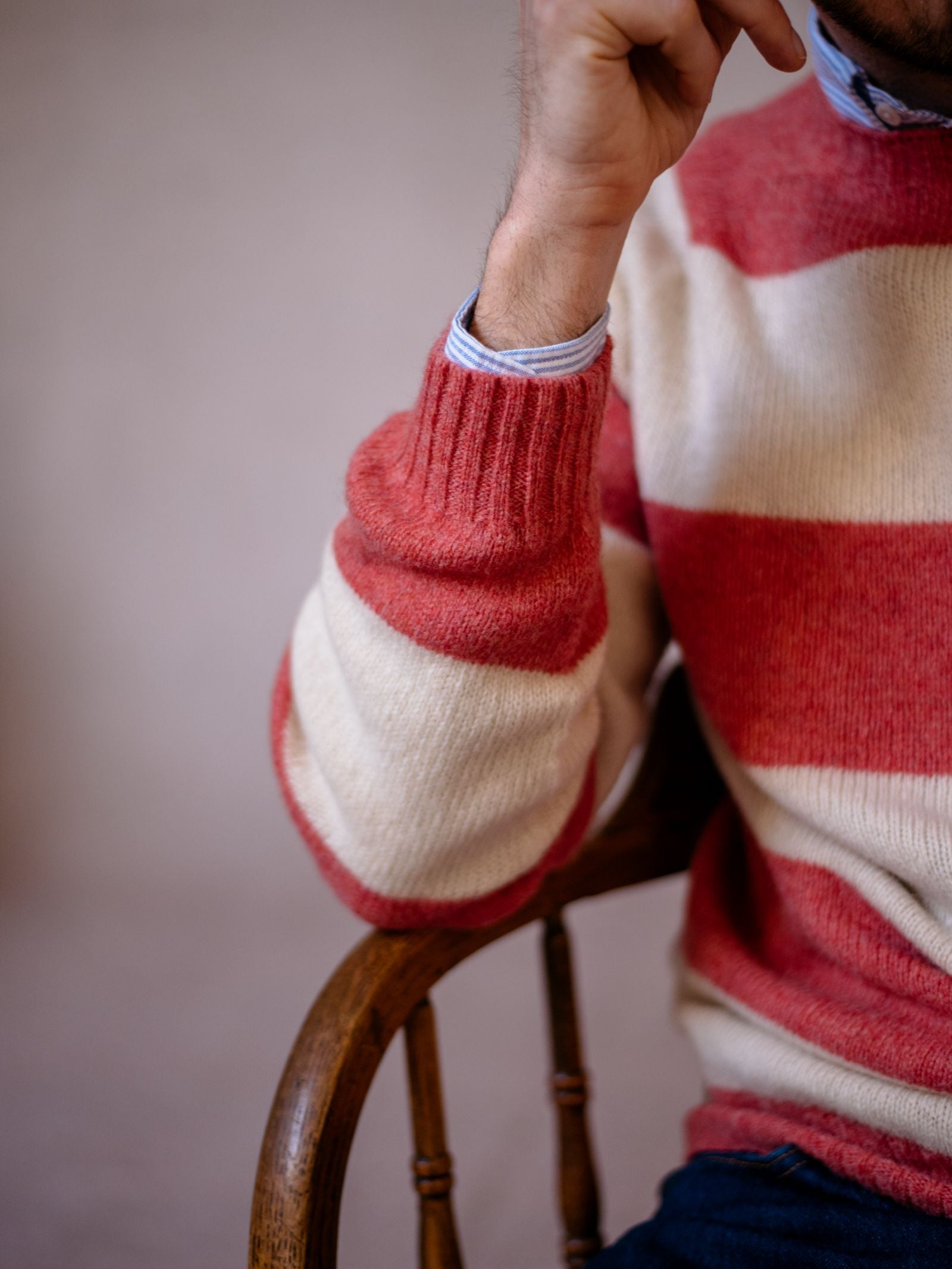 A person wearing the Campbells of Beauly Shetland Stripe Crew Jumper over a blue shirt sits on a wooden chair, resting their chin on their hand. Only their upper body and arm are visible.