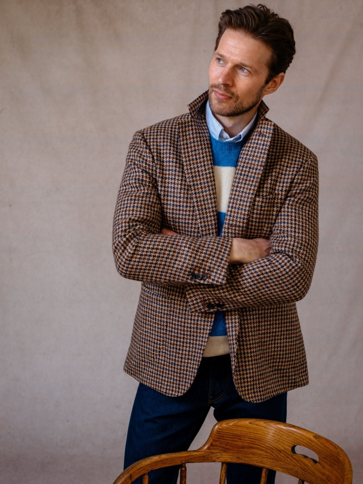 A man with brown hair and a beard stands beside a wooden chair, arms crossed, wearing Campbells of Beaulys Navy Gun Club Tweed Jacket over a striped blue and cream sweater with dark jeans, looking off to the side.