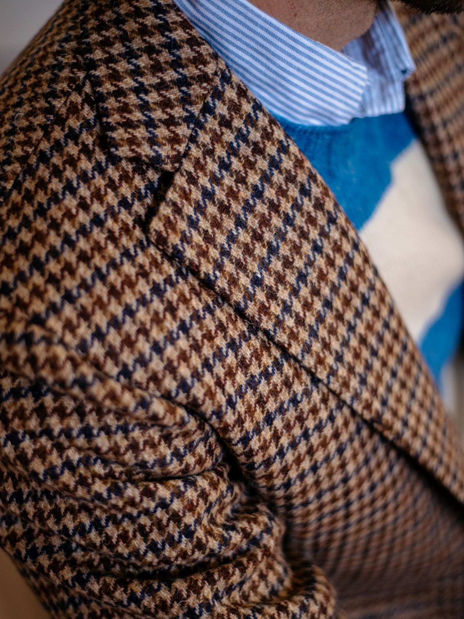 Close-up of a person in the Campbells of Beauly Navy Gun Club Tweed Jacket, featuring a brown and blue houndstooth pattern, worn over a blue and white sweater and a light blue striped shirt; only part of the face and upper torso are visible.