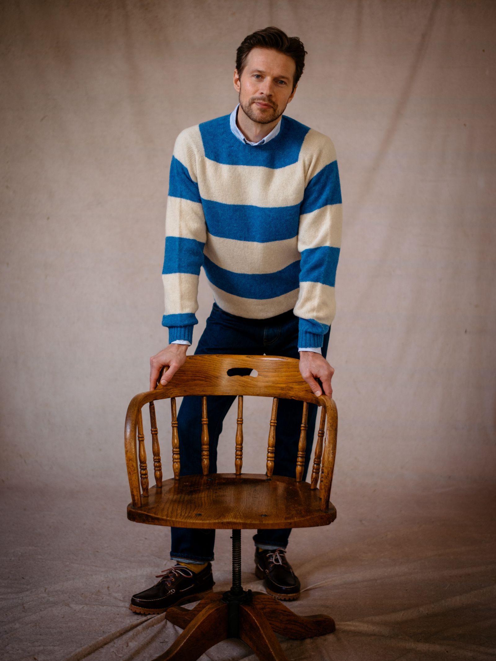 A man with short hair wears the Campbells of Beauly Shetland Stripe Crew Jumper and dark pants, standing behind a wooden chair with his hands on its back, against a neutral indoor backdrop.