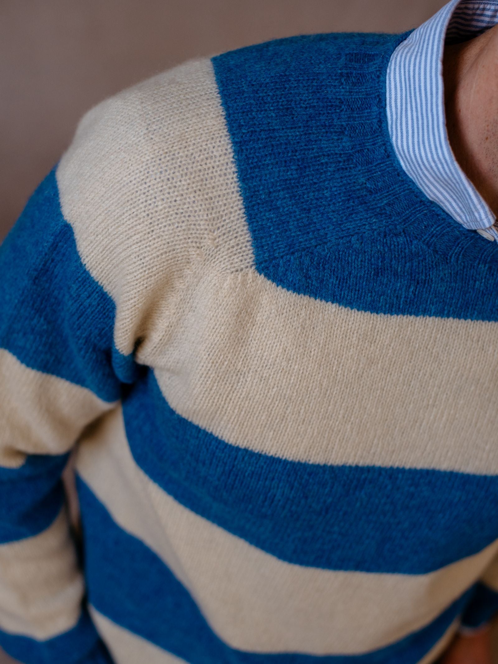 A person wears the Campbells of Beauly Shetland Stripe Crew Jumper, featuring wide blue and beige stripes, over a blue and white striped collared shirt. The photo is taken from above, showing only the upper torso and part of the neck.