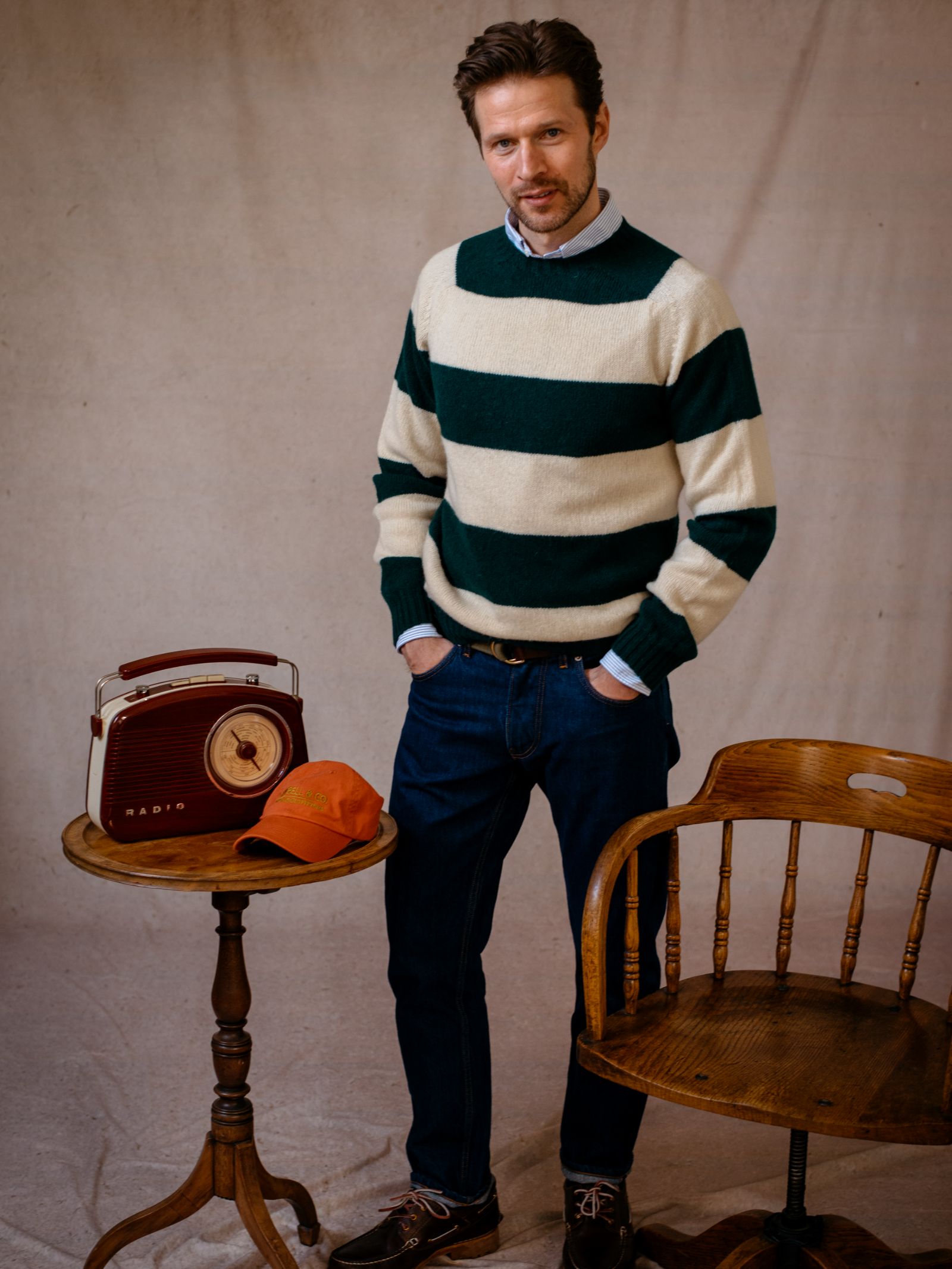 A man in the Campbells of Beauly Shetland Stripe Crew Jumper and jeans stands indoors by a small table with a vintage radio and orange cap, with a wooden chair close by.