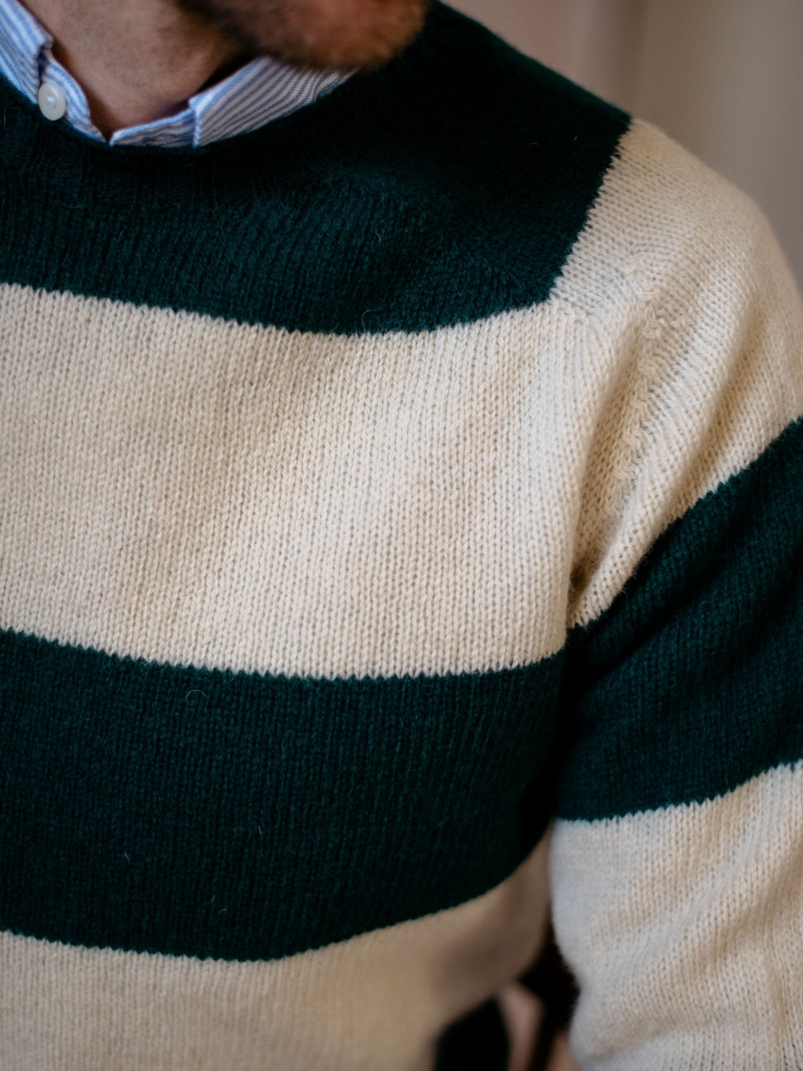 A close-up of a person wearing the Campbells of Beauly Shetland Stripe Crew Jumper, featuring wide dark green and cream stripes, layered over a blue and white collared shirt. Only part of their chest and shoulder is visible.