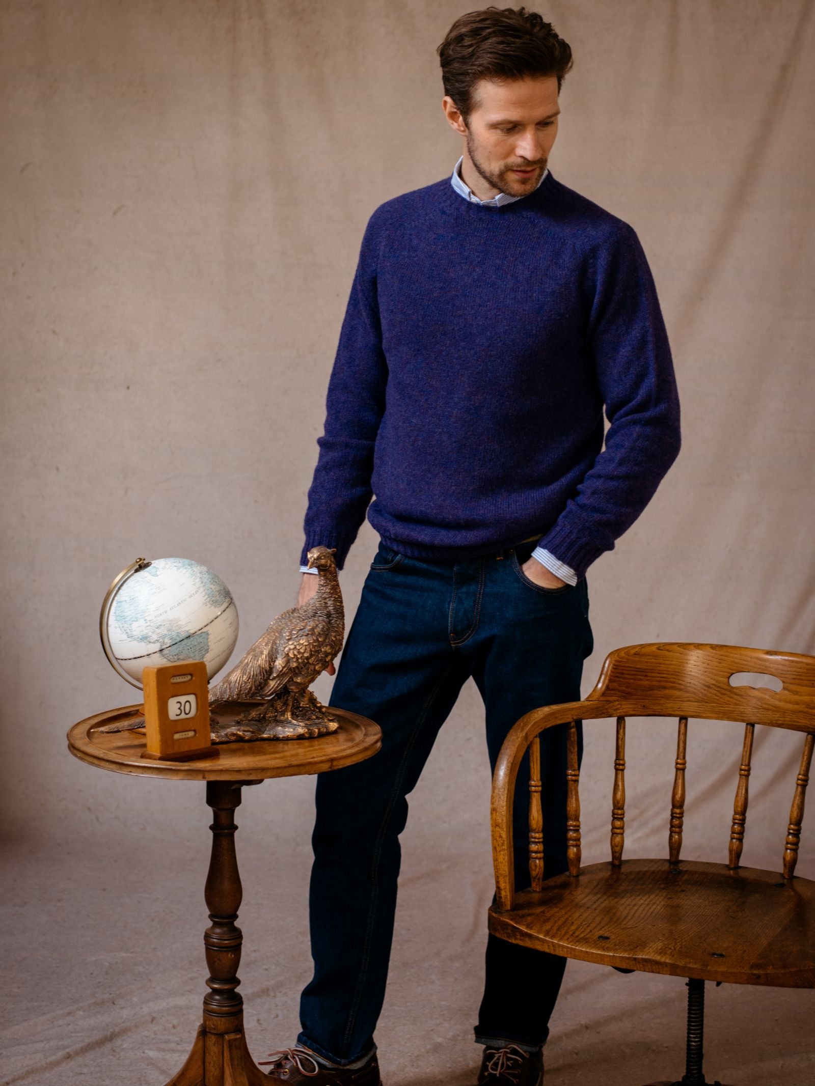 A man wears the Campbells of Beauly Shetland Jumper, made from lambswool, with jeans. He stands by a small round table holding a globe, bird statue, and calendar. A wooden chair is nearby; the backdrop is plain beige fabric.